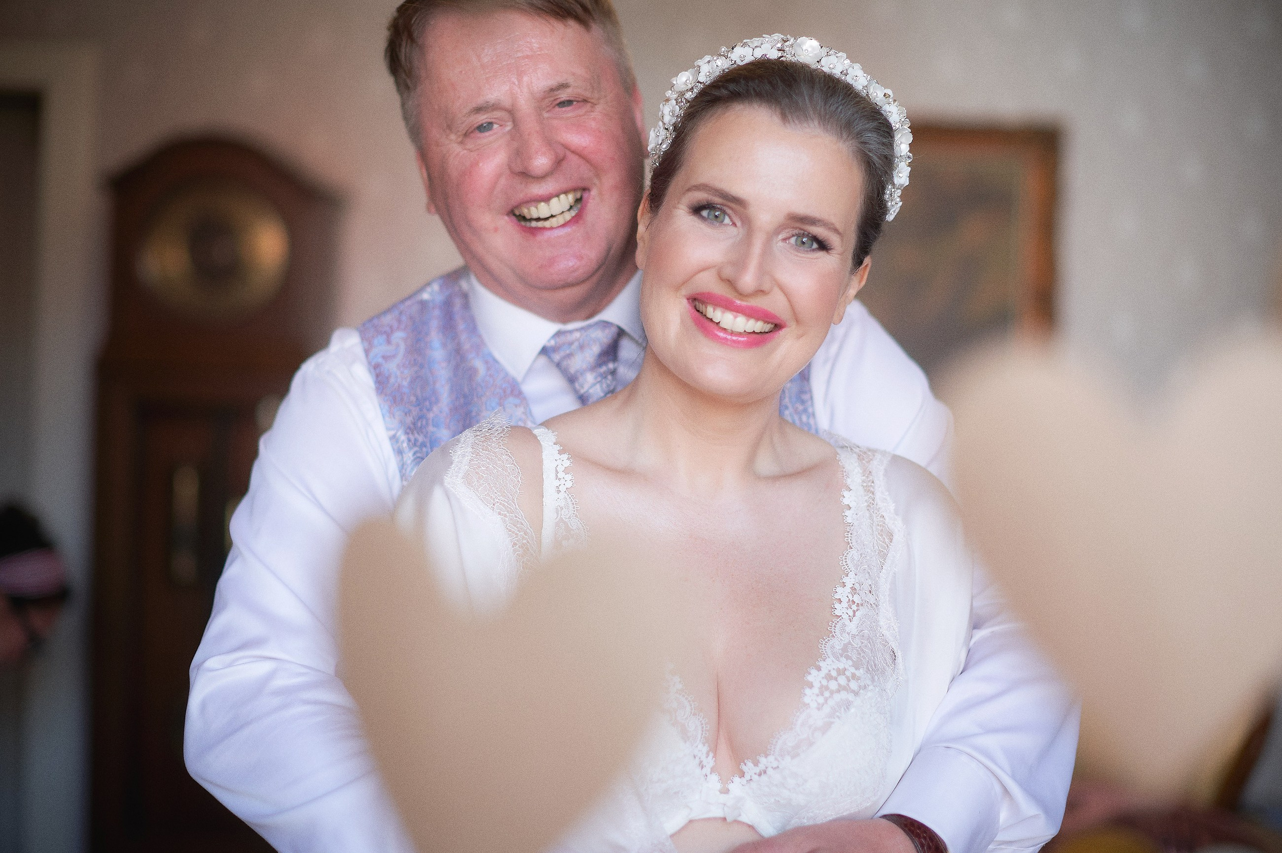 A brunette bride wearing a tiara smiles as she is hugged from behind by her father during her wedding day preparation at the Hotel Constance in Prague.