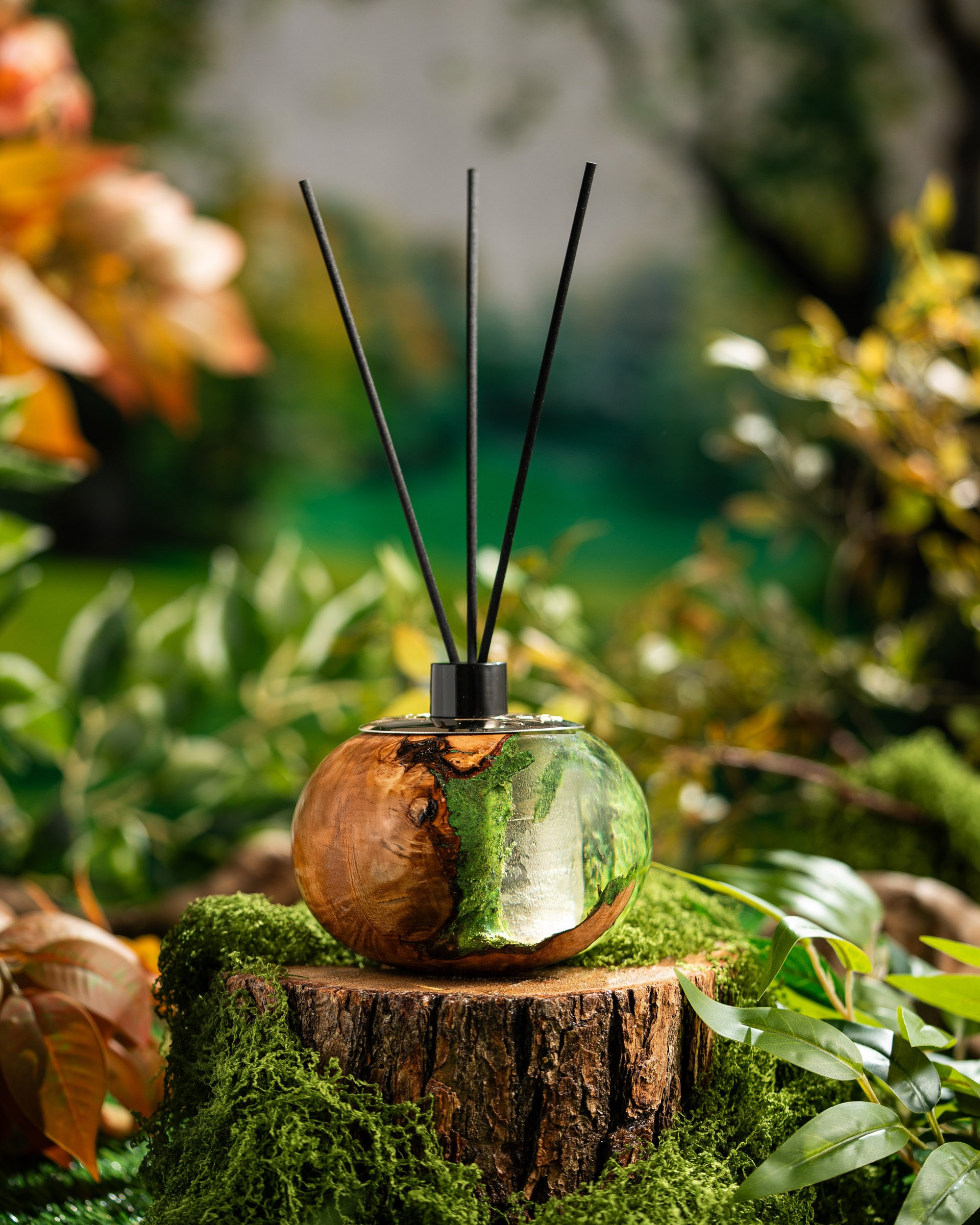 Ağac və yaşıl qatran dizaynlı, qara çubuqları olan otaq qoxusu diffuzoru gövdənin üstündə, yosunlu və yaşıllıqla dolu meşə fonunda təqdim olunur.