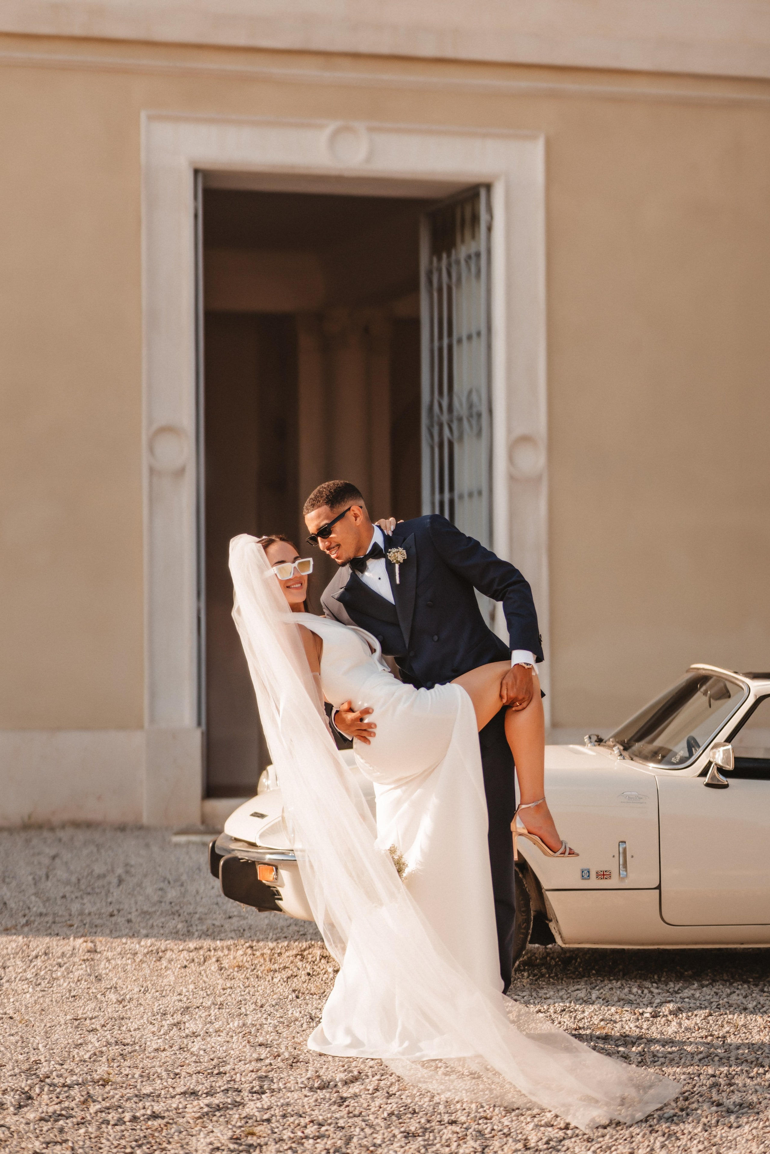 Noivo carrega a noiva com romantismo em frente a carro vintage numa cerimónia sofisticada.