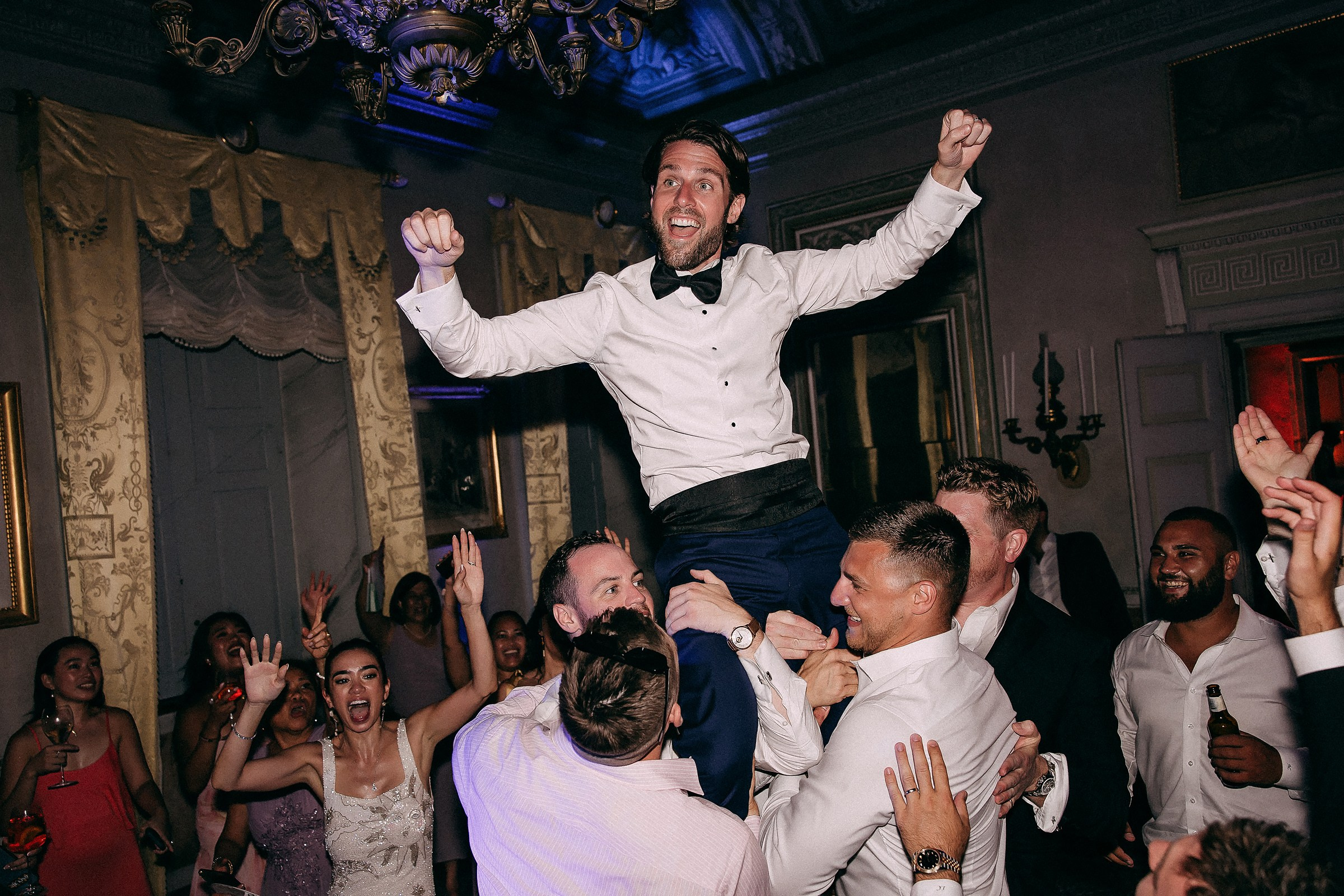Groom raised high on friends’ shoulders, celebrating with joy and laughter on the dance floor.
