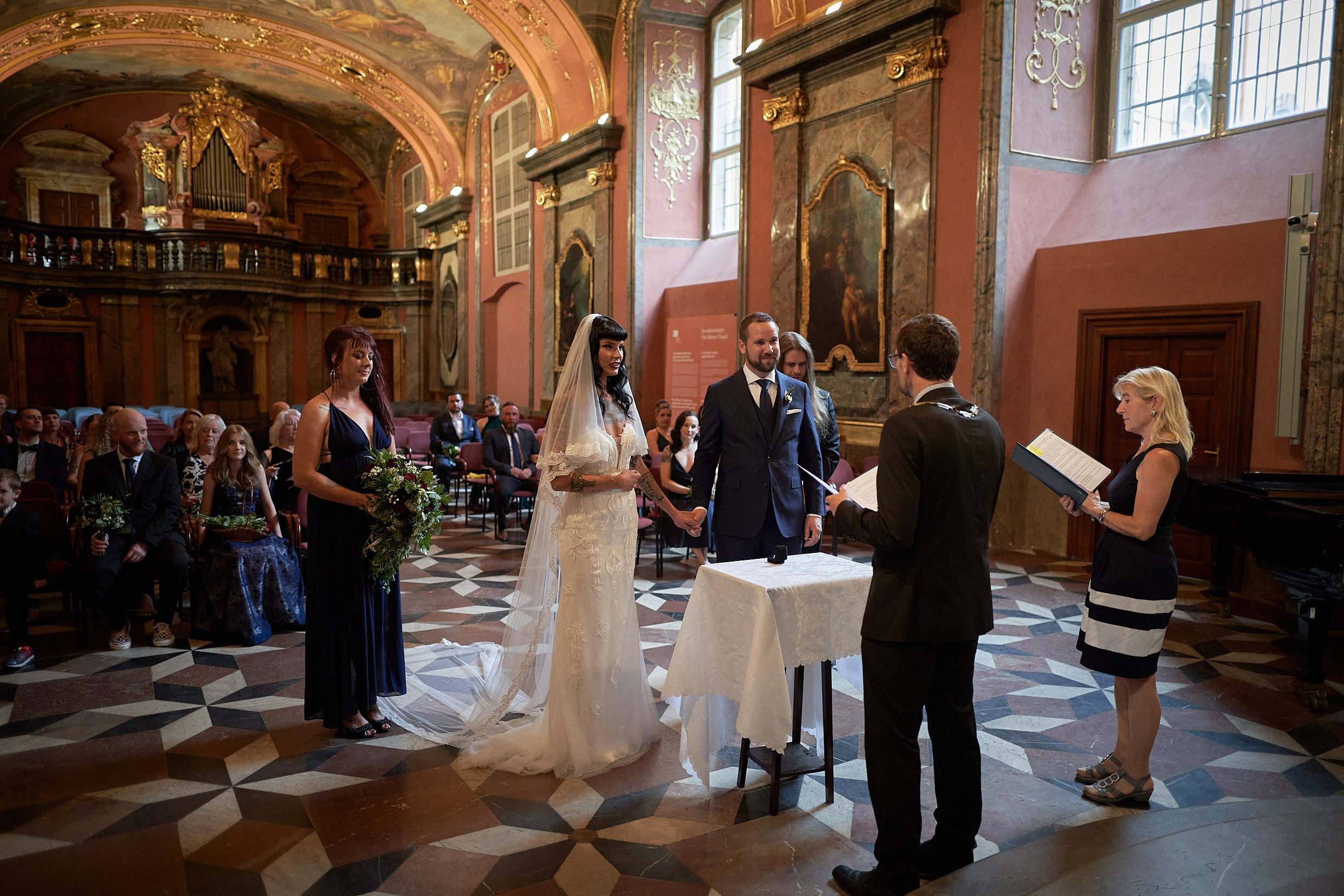 Newlyweds holding hands listening to officiant in baroque chapel.