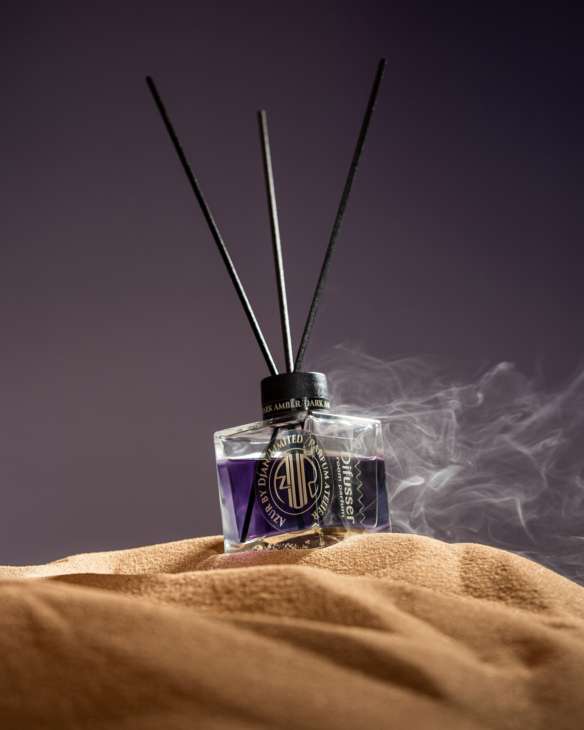 AZUR by DJAN brendinə aid, bənövşəyi maye ilə doldurulmuş kvadrat formalı şüşəli qamış diffuzor (ətir yayıcı) açıq qəhvəyi qum yığınının üzərində yerləşdirilib. Şüşədən yuxarıya doğru üç qara qamış çıxır. Şüşənin sağ tərəfindən nazik bir tüstü və ya ətir dumanı qalxır. Arxa fon sadə, tünd bənövşəyi-bozdur.