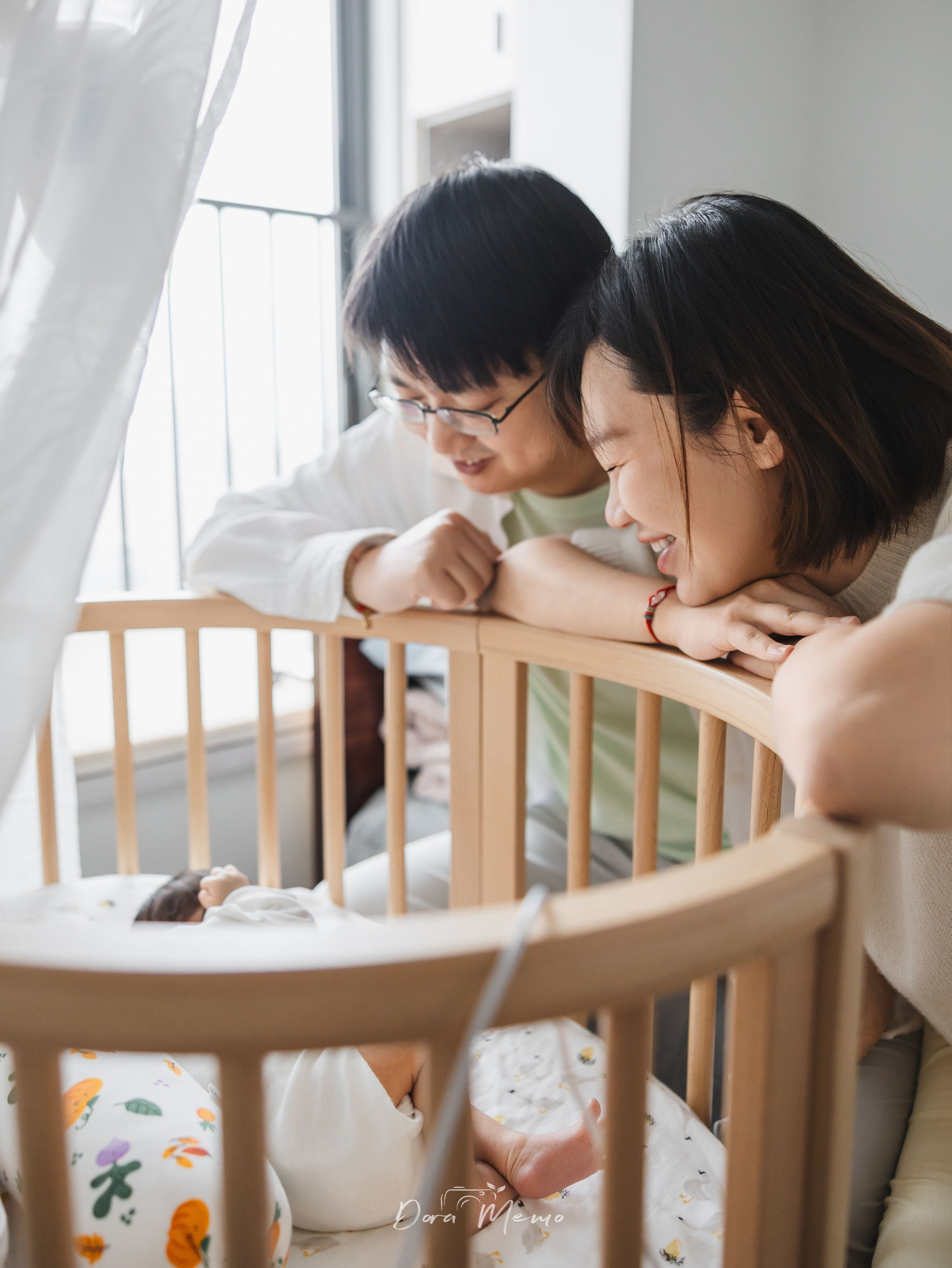 New parents leaning over a crib together, gazing at their newborn baby in a bright Shanghai apartment