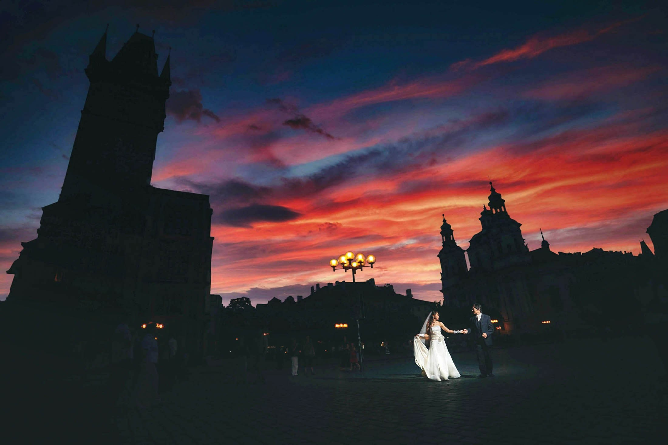 Newlyweds in Old Town Square stand in stark contrast to the darkened square as the sky flares above