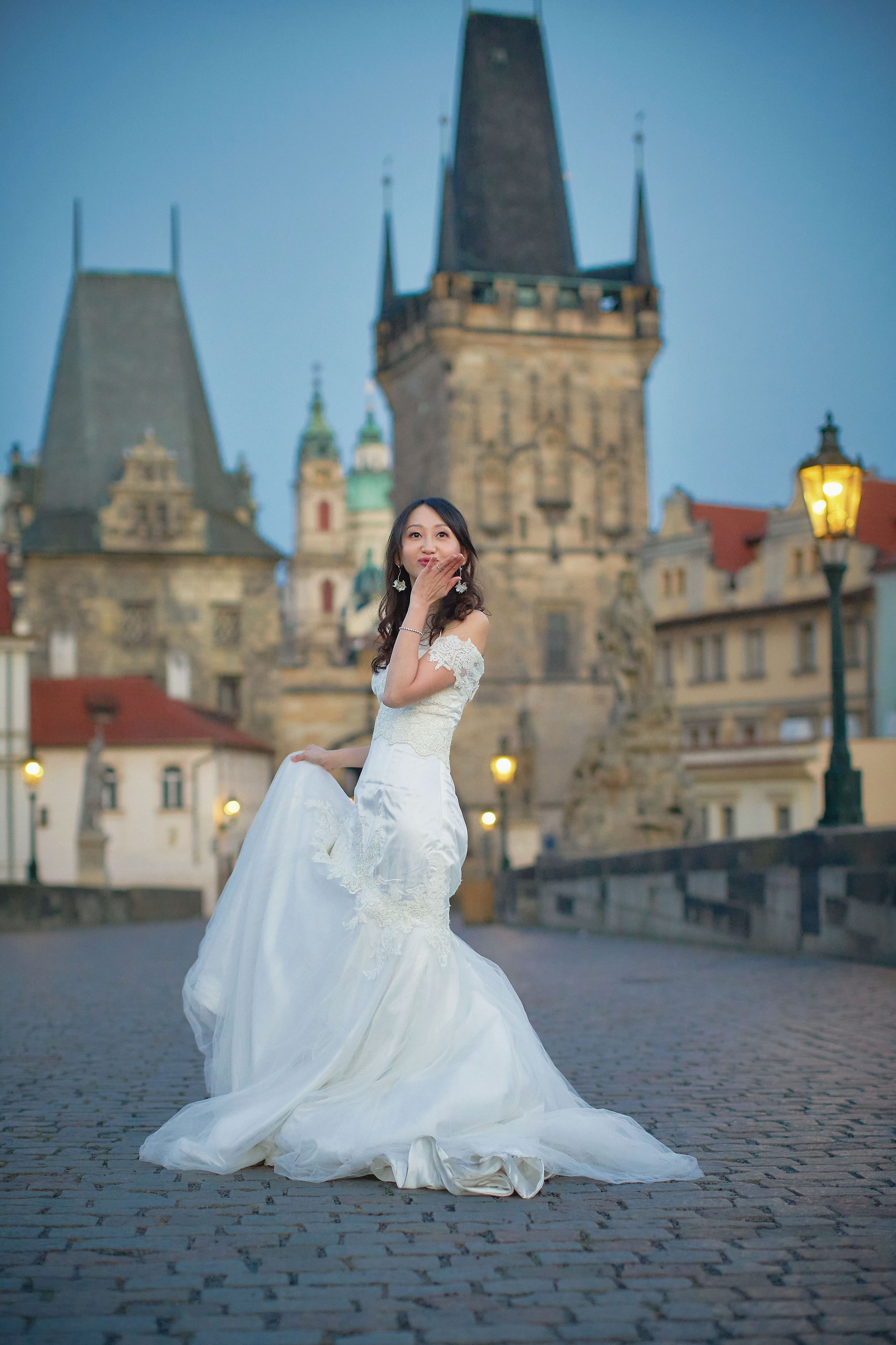 Bride Eva blowing a playful kiss while holding wedding dress on Charles Bridge at dawn.