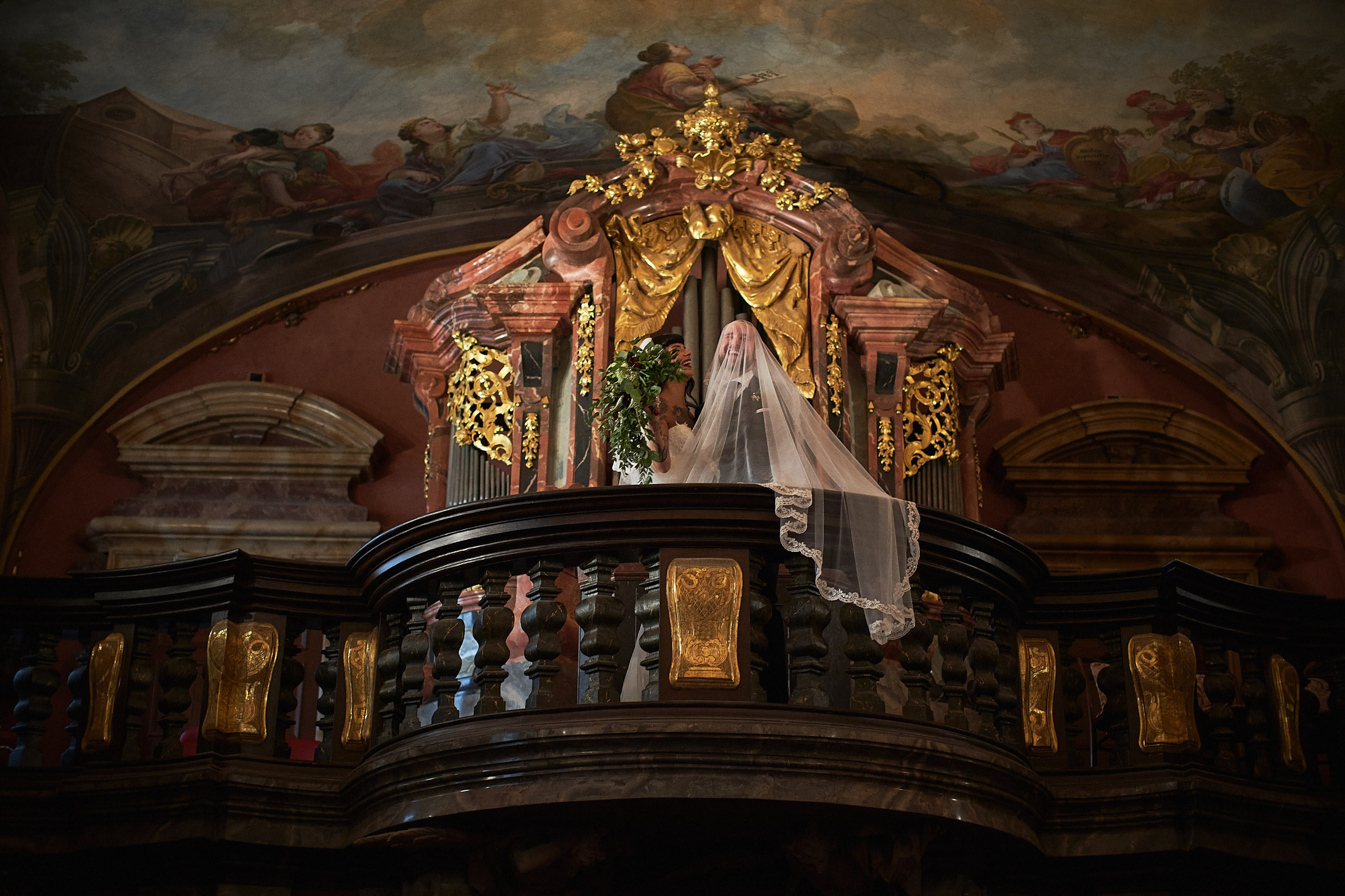 Tattooed bride covering groom's face with veil on chapel balcony.