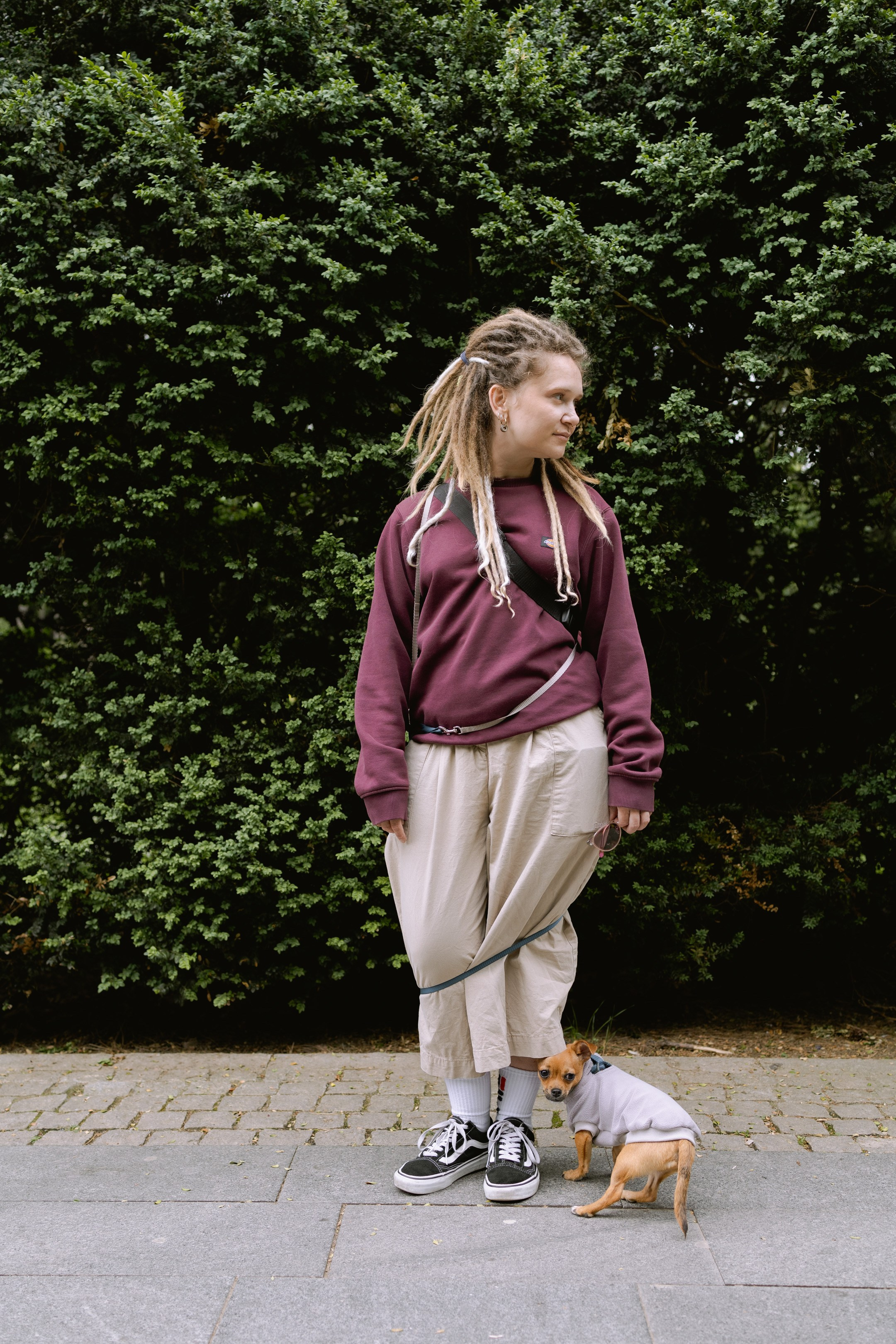 Юля с дредами стоит в парке, а её собака Роки обвила её поводком.