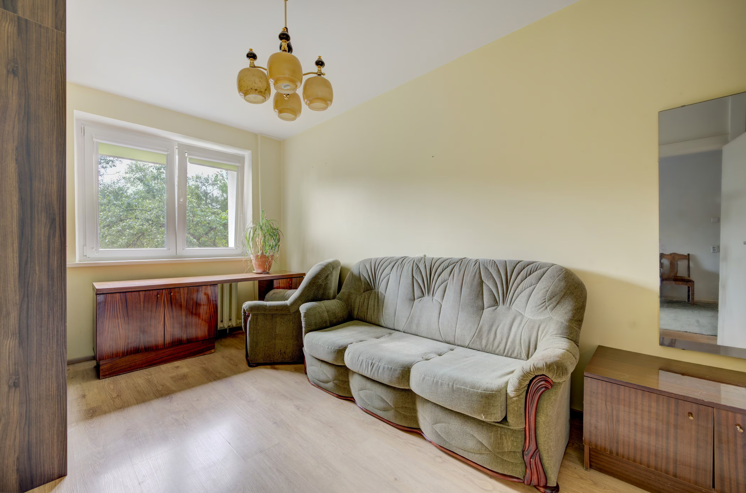 Jaukus gyvenamojo kambario interjeras su šviesia sofa, krėslu, medine spintele ir šviestuvu.