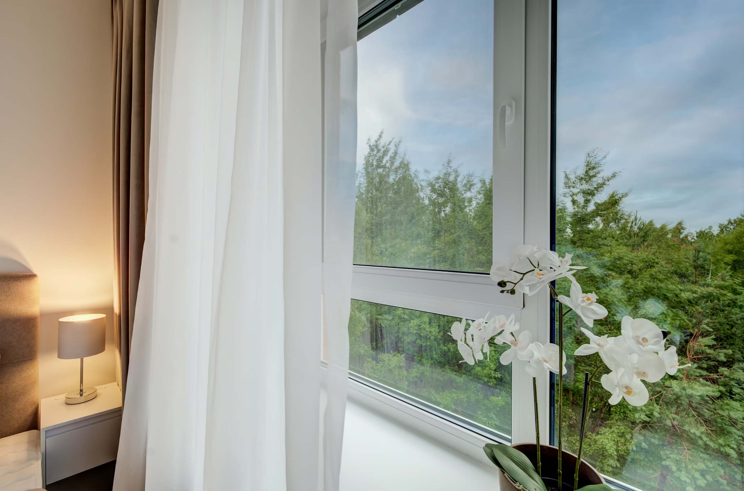 Modernus miegamojo langas su perregimomis užuolaidomis, orchidėja ir vaizdu į gamtą.
