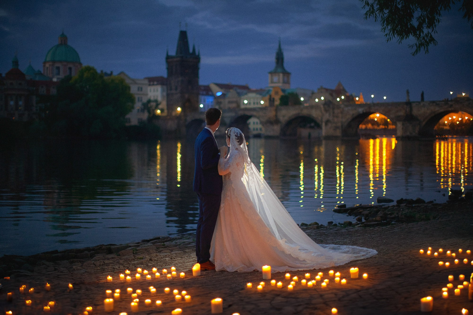 As the gas lamps illuminate the historic Charles Bridge in the background, a bride and groom stand surrounded by candles as they await the sunrise in Prague.