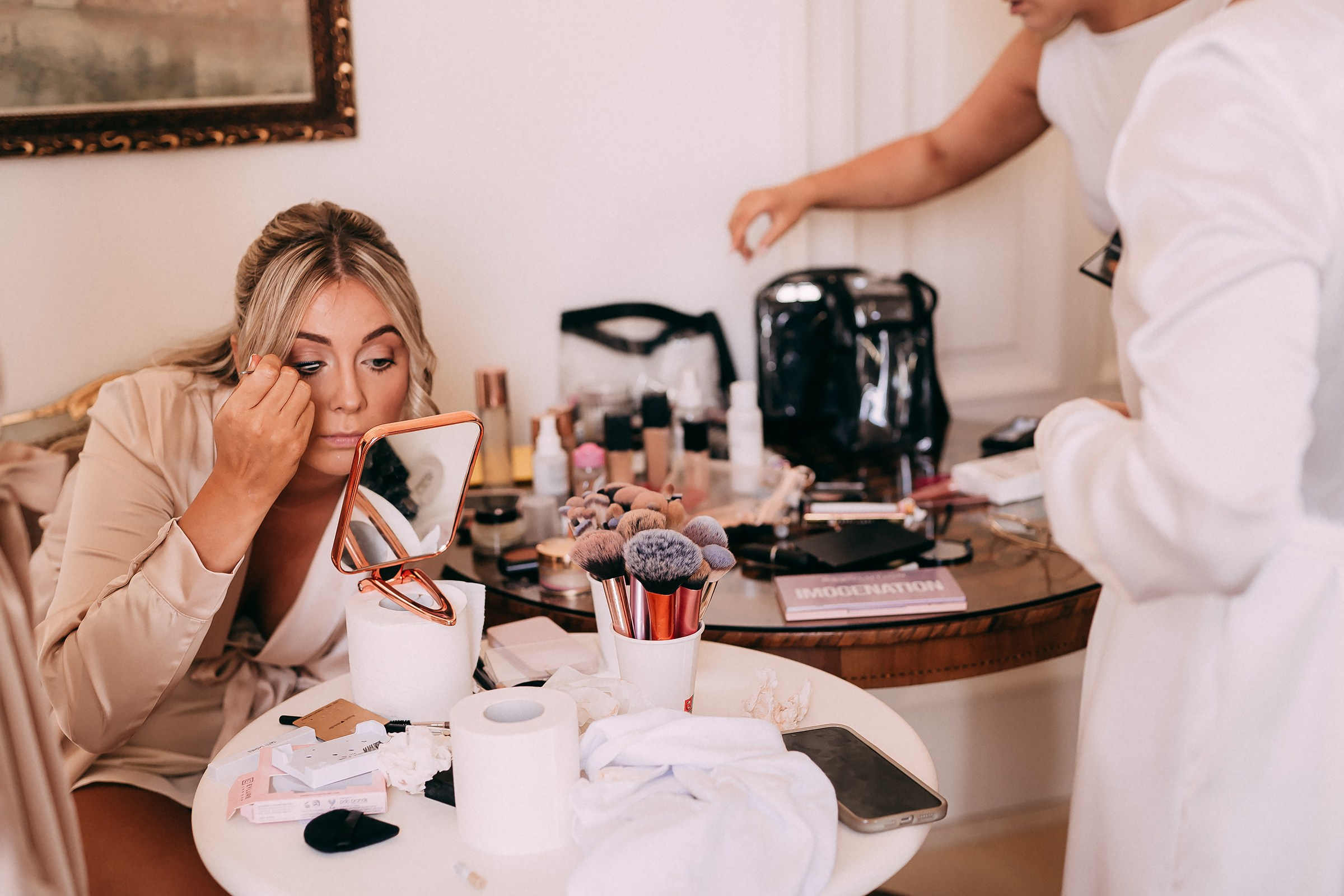 A bridesmaid leans in close to a mirror, carefully applying makeup at a preparation station filled with beauty tools inside a serene bridal suite.
