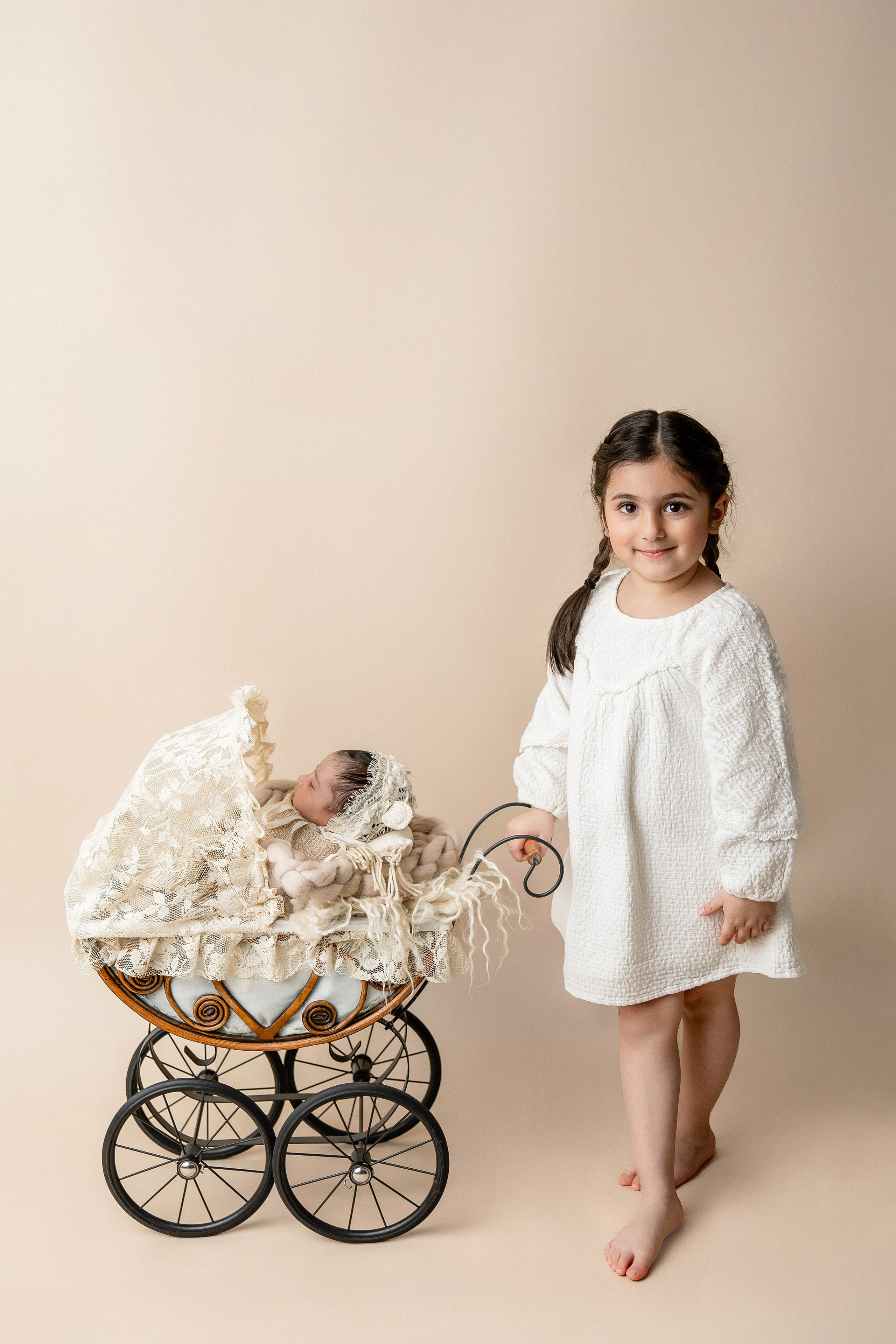 Big sister in a white dress standing beside a vintage stroller with her newborn sibling sleeping inside — professional sibling and newborn photography on neutral background.