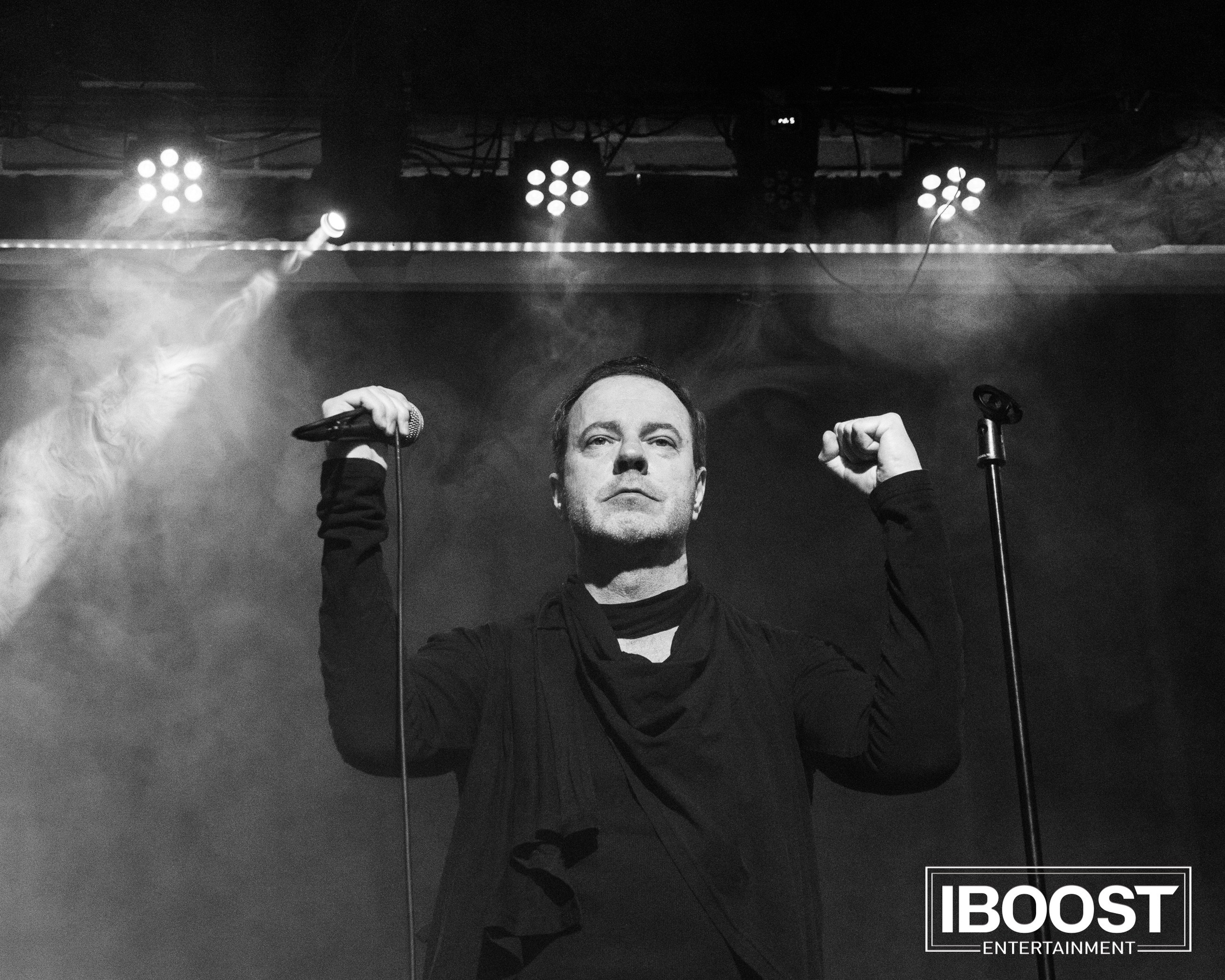 Black-and-white stage photo of the lead vocalist of Animal JazZ standing under concert lights with both hands raised.