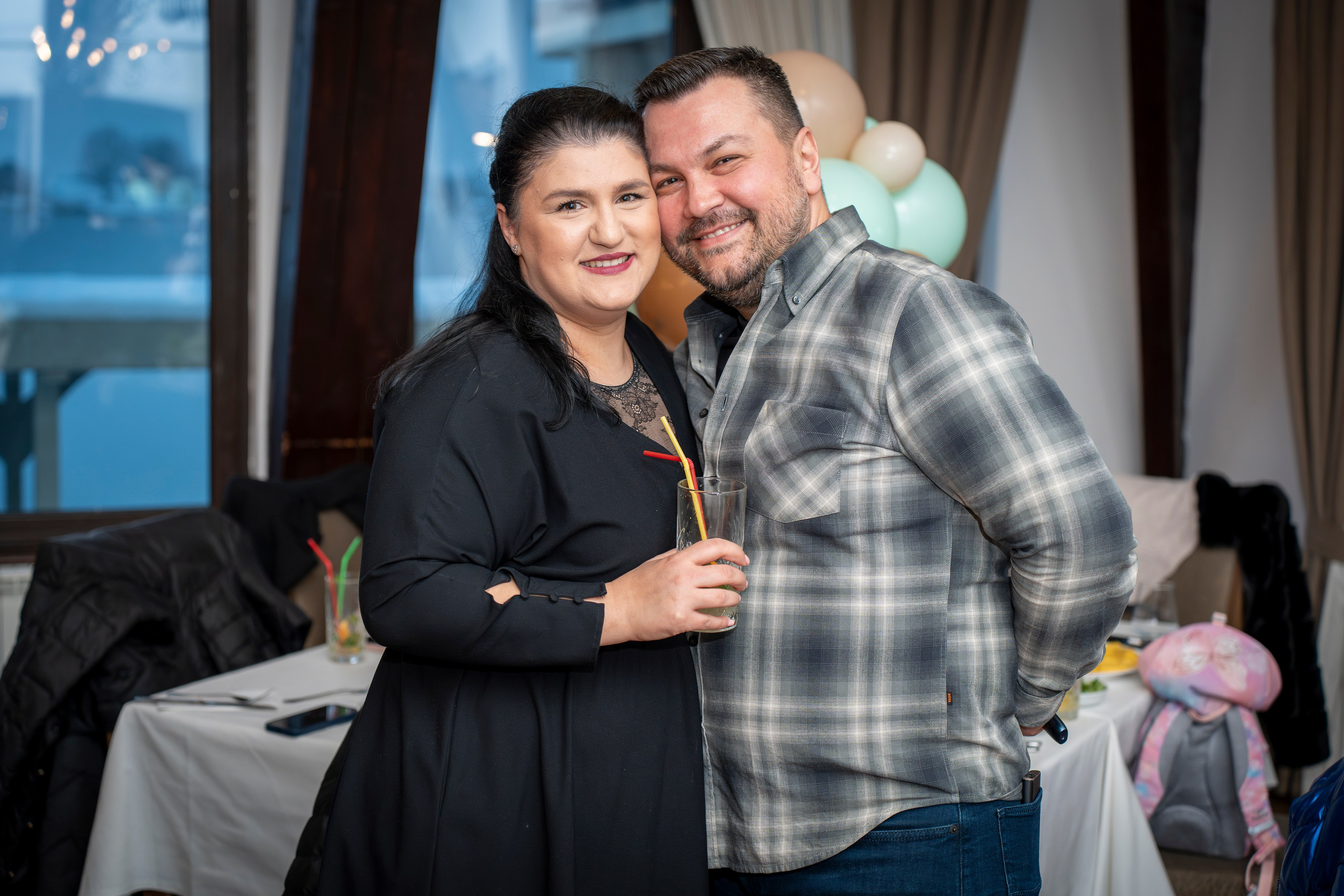 Smiling couple posing indoors near a table with food and balloons in the background.