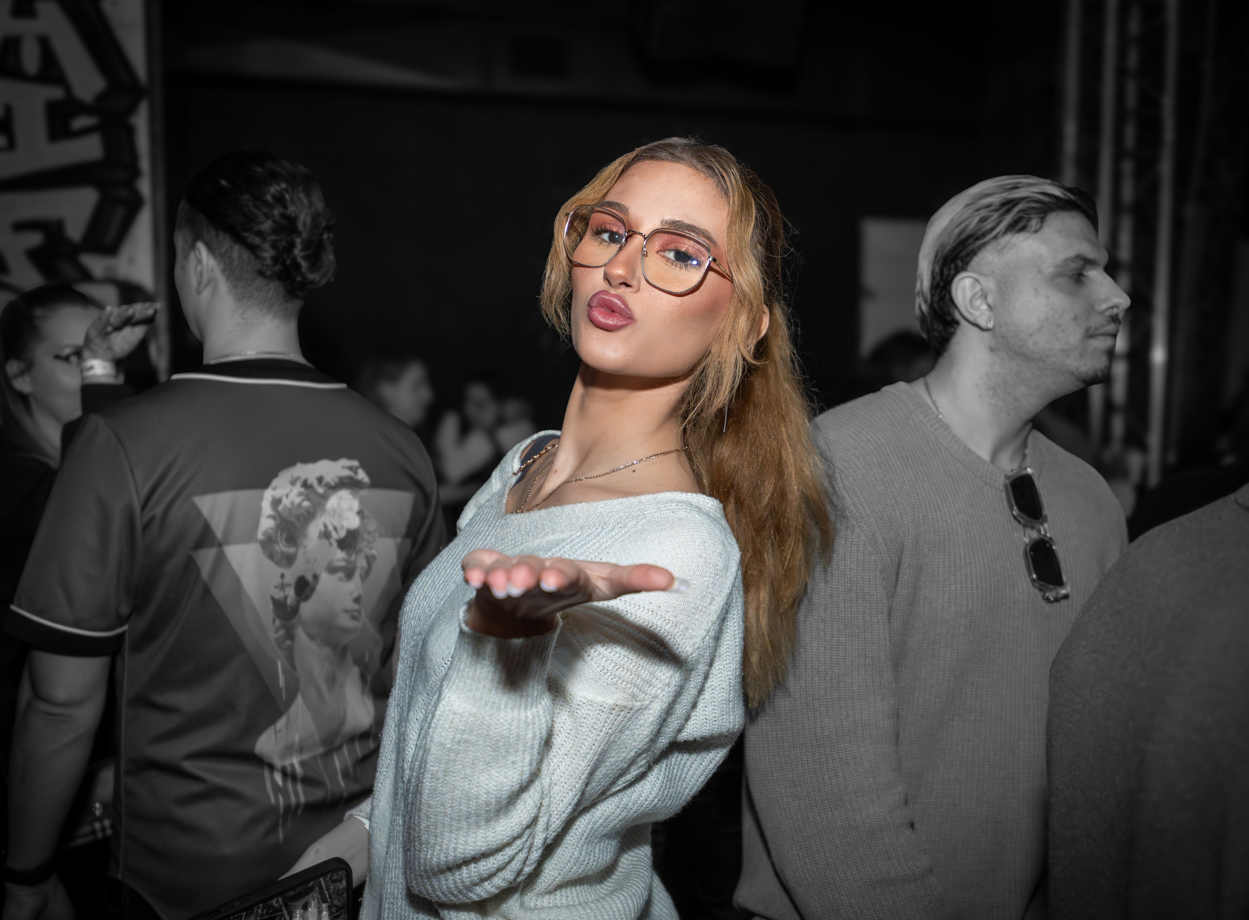 Young woman wearing glasses blowing a kiss at a social gathering at night.