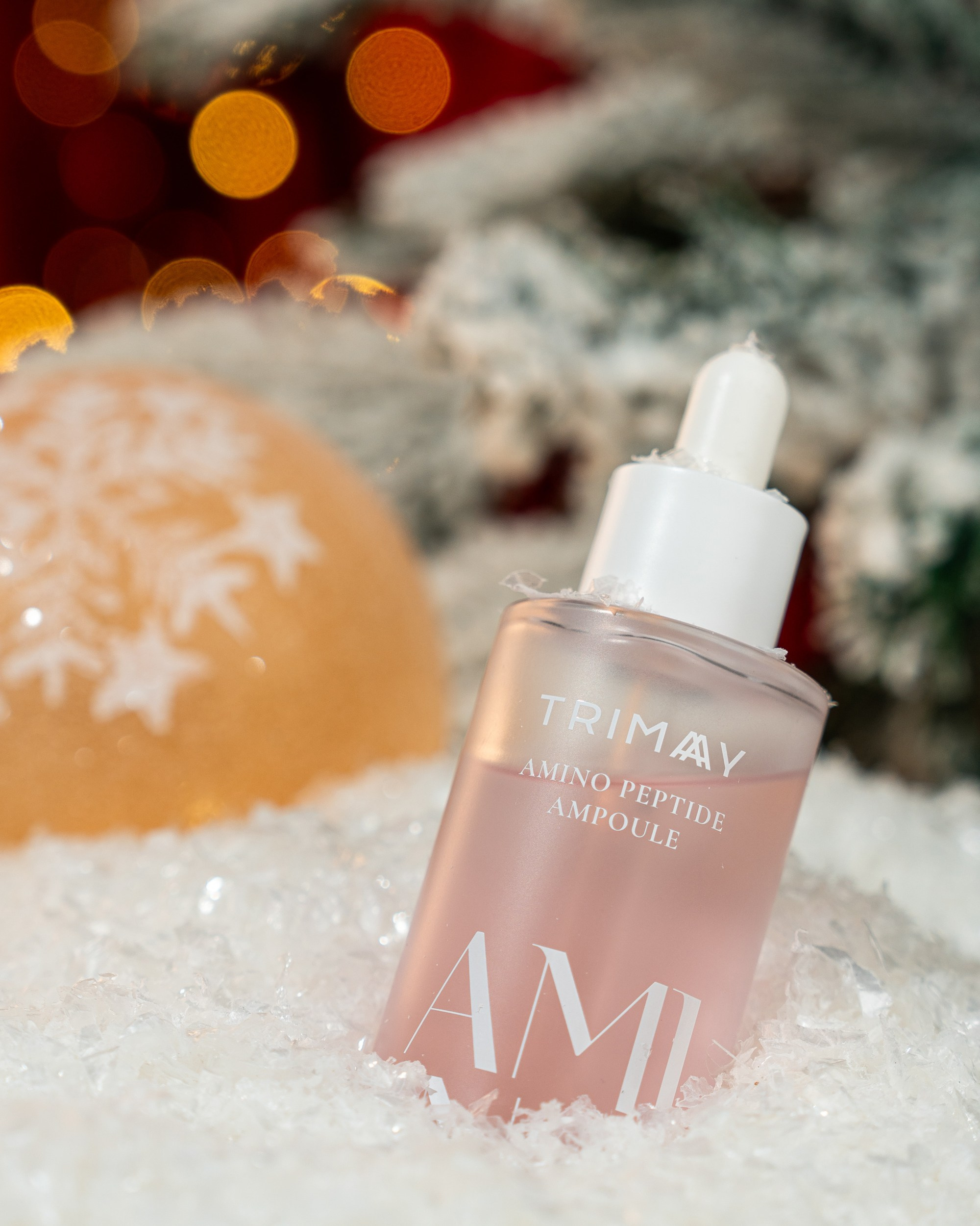 Süni qar və buz parçaları arasında, çəhrayı maye ilə dolu Trimay Amino Peptid Ampula butulkası. Arxada böyük, portağalı rəngli qar dənəciyi bəzəyi və bulanıq işıqlar görünür.