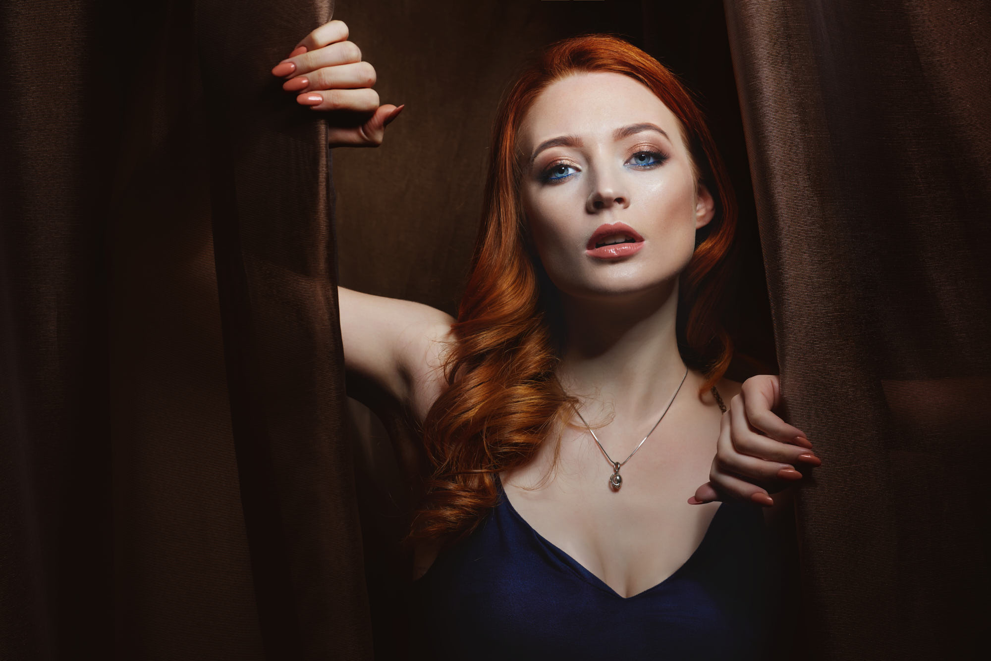 Stunning beauty portrait of a red-haired model wearing a necklace, emerging from behind dark curtains. Perfect for jewelry advertising.