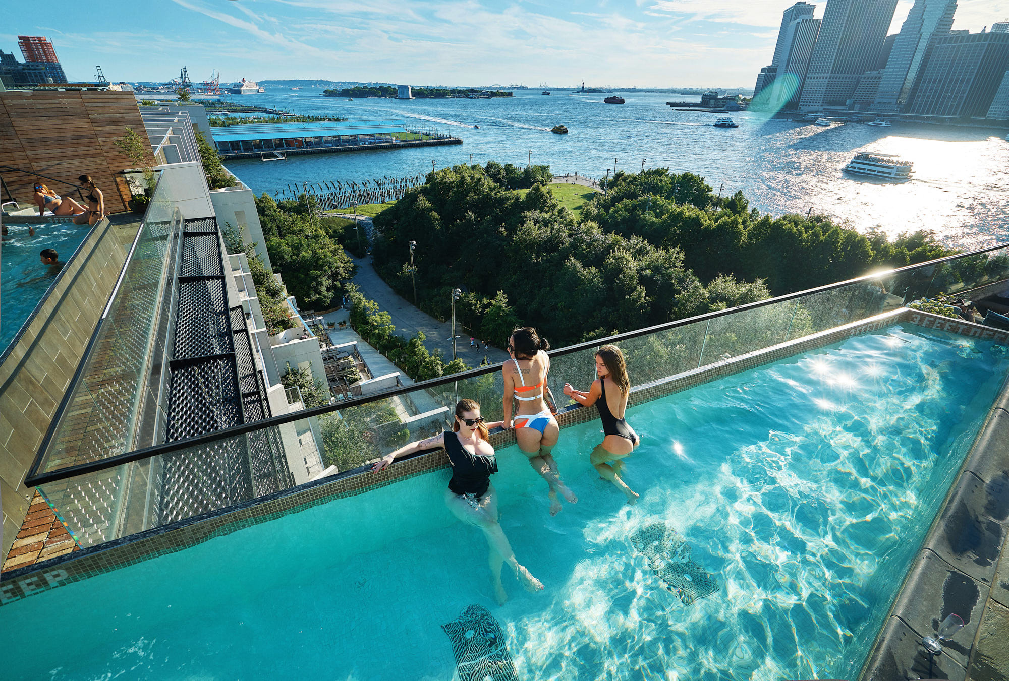 wide angle shot oirls party in a swimsuits in a pool photoshoot on the rooftop pool of the One Hotel Brooklyn Bridge with the Manhattan skyline in the background.