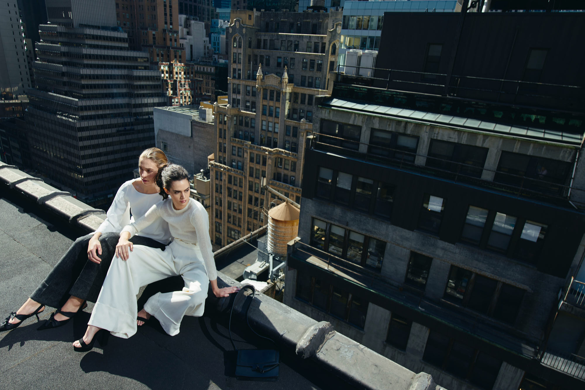 Two women sitting on the edge of a rooftop with the New York City skyscrapers in the background. An editorial fashion commercial photoshoot.