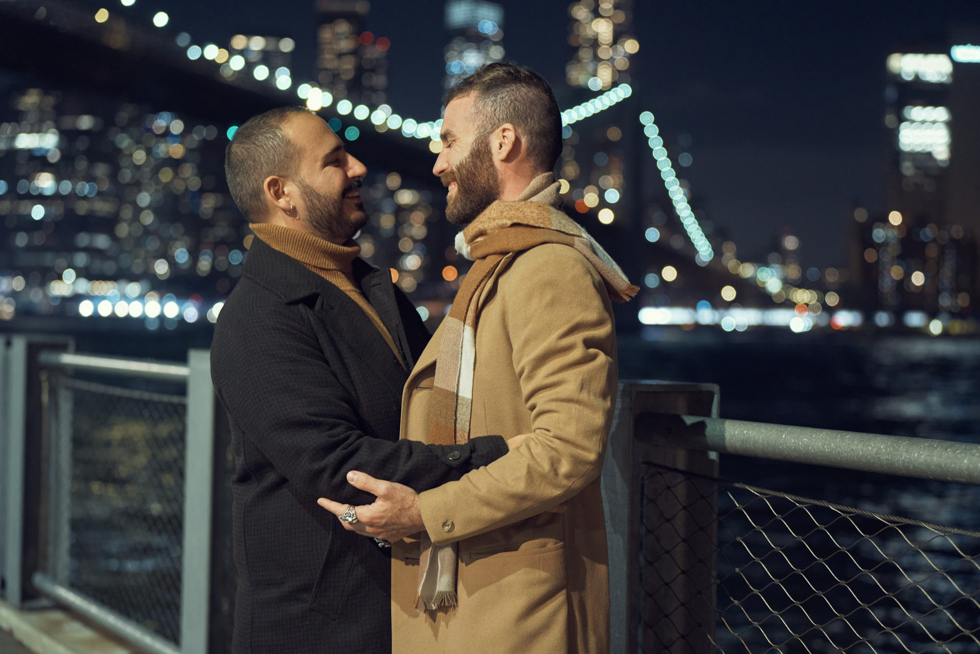 Night engagement photo of gay couple kissing in Dumbo with iconic view on Brooklyn Bridge and Manhattan.