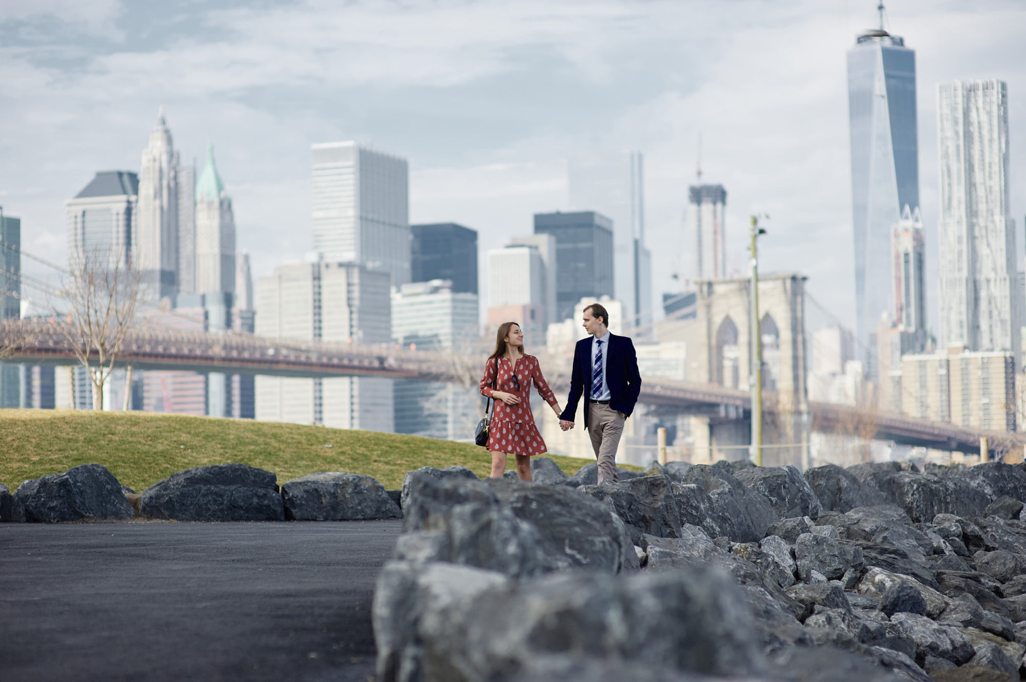 A romantic couple's photoshoot capturing moments of love and connection as they walk along the riverside in Dumbo, with the iconic view of the Brooklyn Bridge and Manhattan skyline as a backdrop.