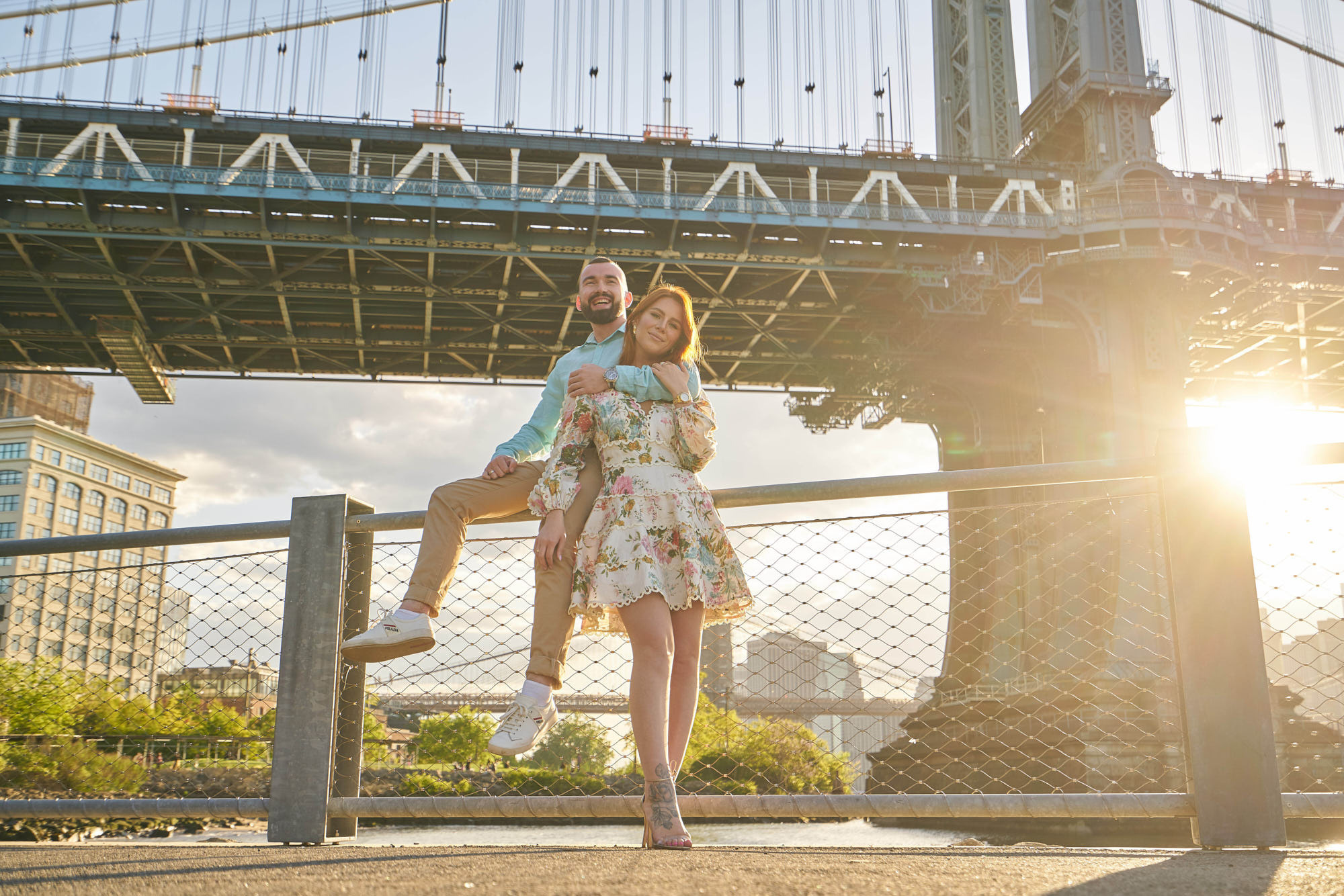 Romantic evening engagement photoshoot under Brooklyn Bridge capturing playful moments, with the groom-to-be jumping high while gazing into his partner's eyes.