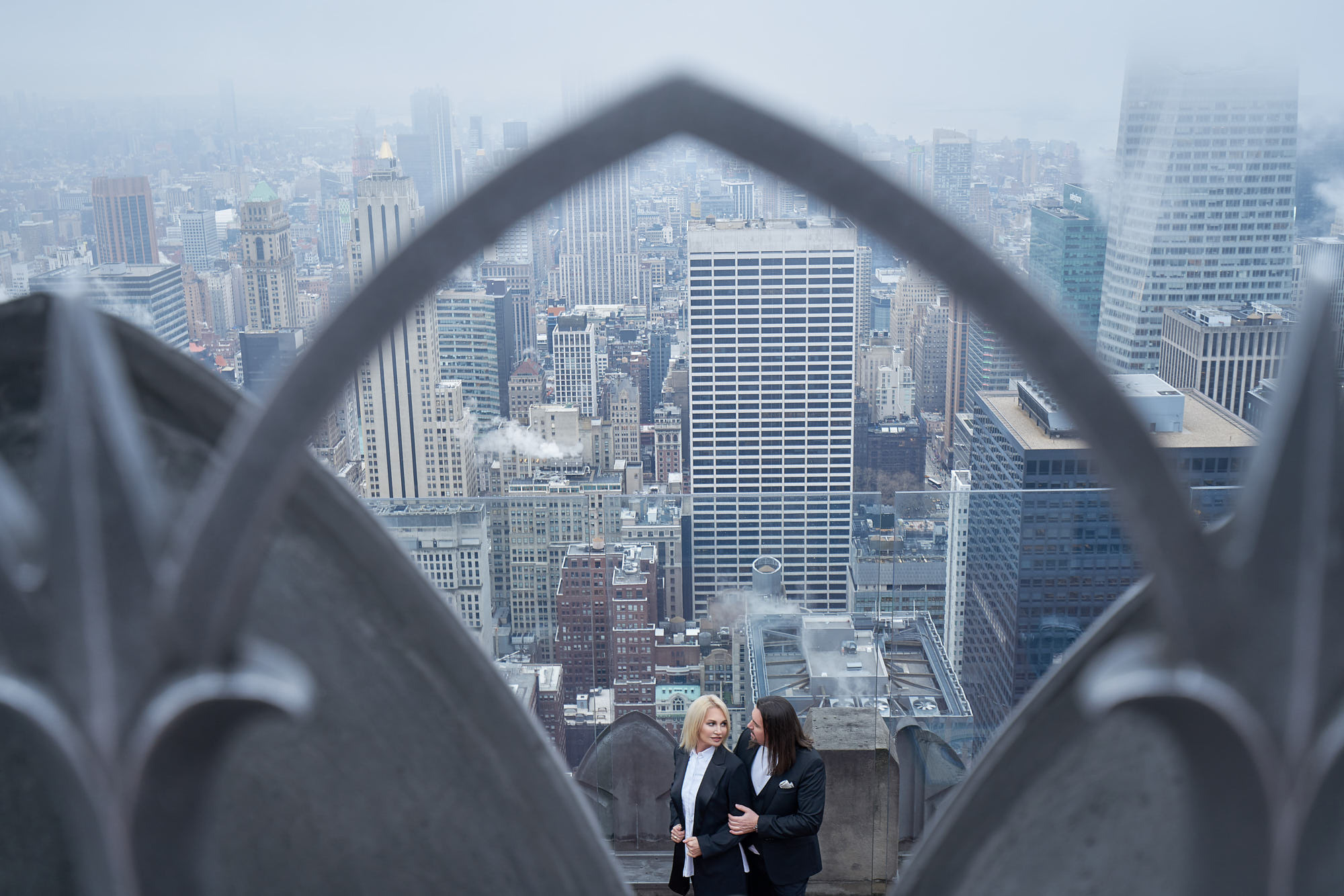 "Top of the Rock" engagement photo featuring Alex and Tatyana in black suits hugging with breathtaking Manhattan skyline view.