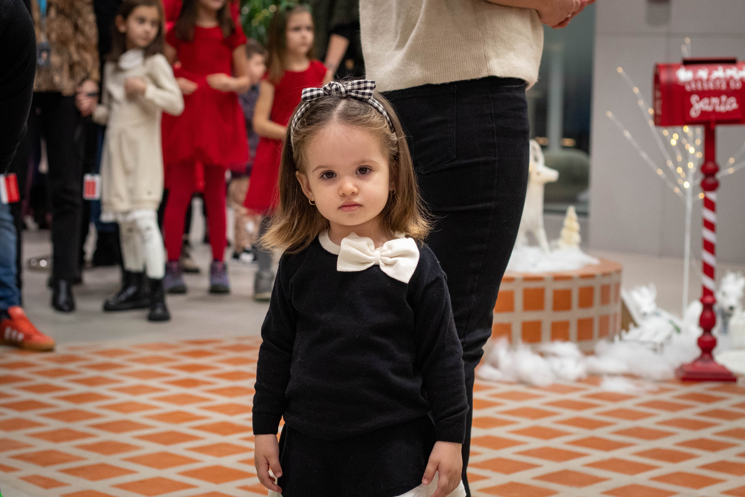 Little girl in black dress with white bow standing indoors at a Christmas event.