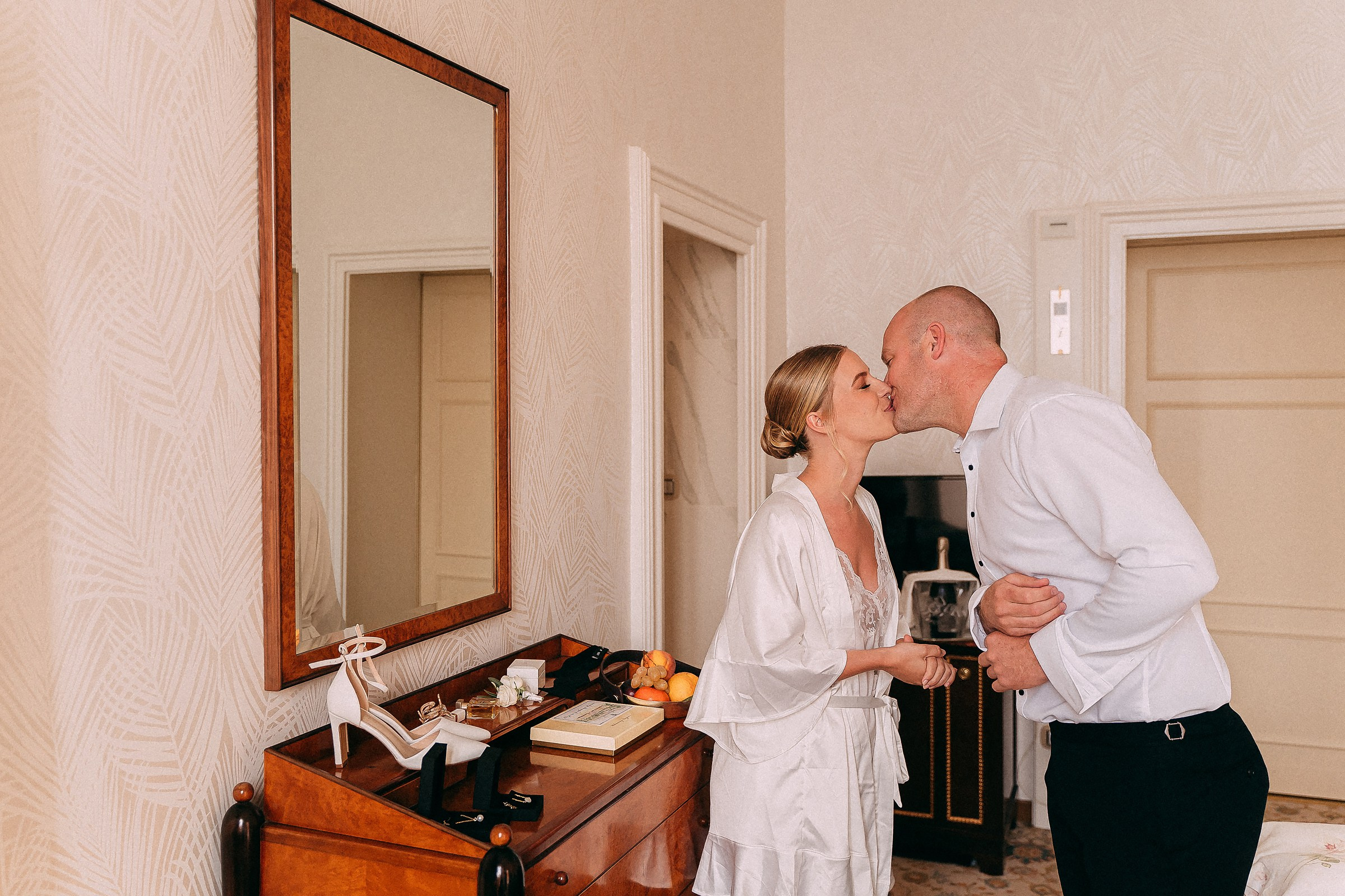 An intimate and joyful moment as a bride shares a kiss with a loved one in a warmly lit room, with wedding accessories arranged on a wooden dresser.