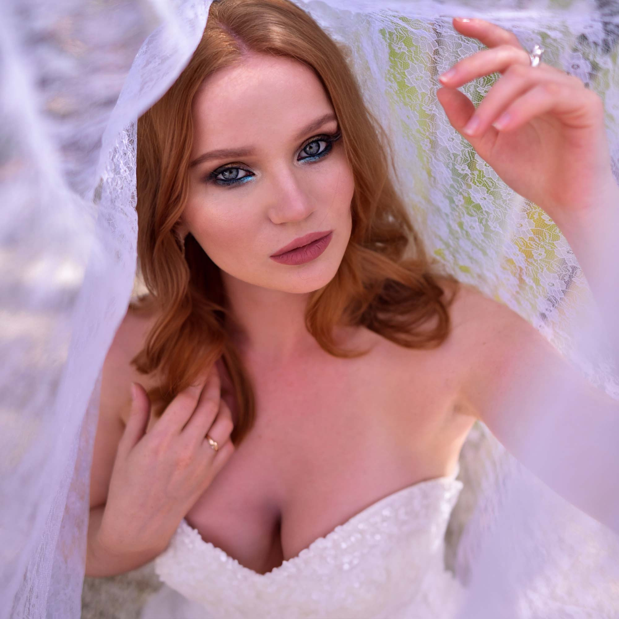 red haired woman with intense smoky eye makeup with a blue accent in a wedding dress ander a white veil