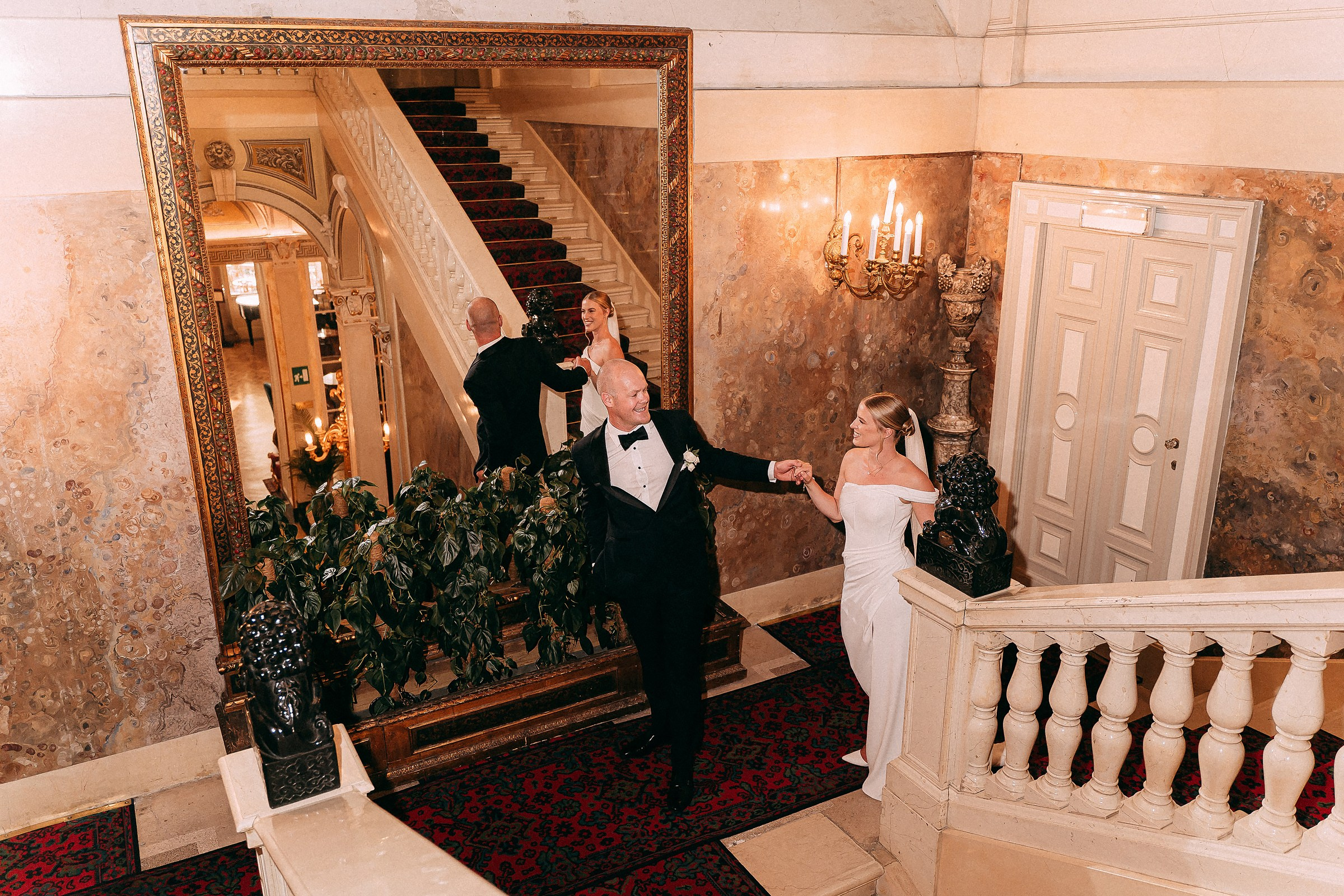 The bride and groom, smiling and holding hands, walk down an intricately decorated staircase surrounded by classical architecture.