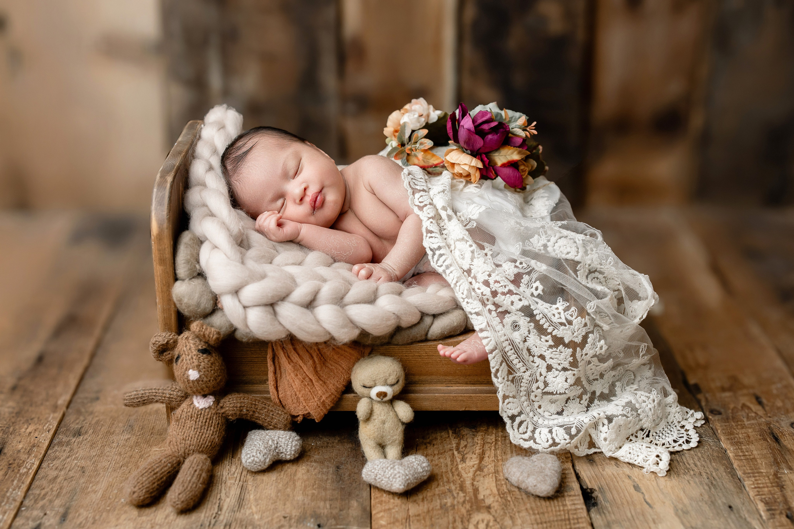 Newborn baby girl sleeping on a miniature bed with lace blanket, surrounded by soft toys and rustic wood background — styled vintage newborn photography setup.