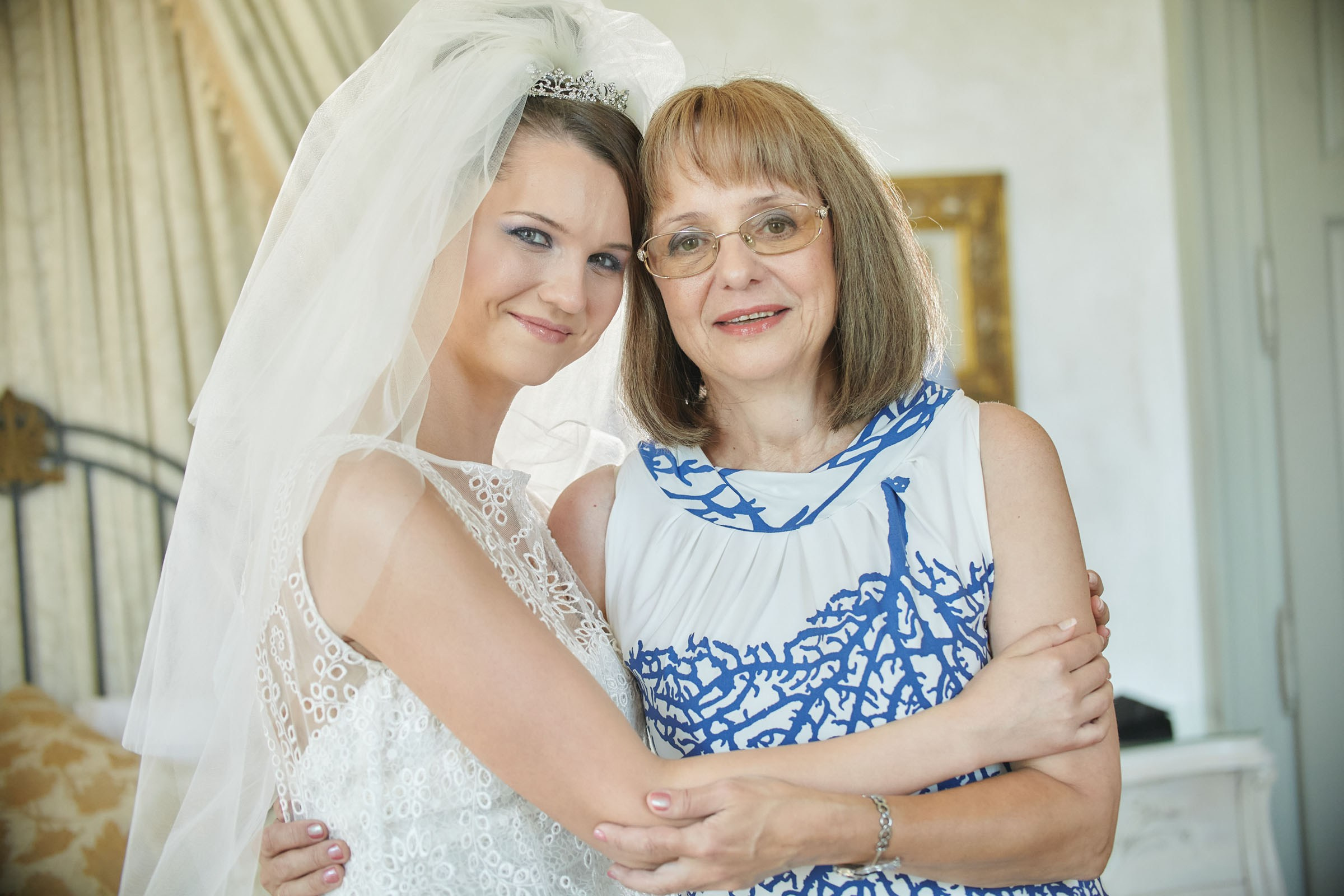 A smiling bride clutches her mother's arms as they both pose for a photo in the bridal suite of the Chateau Mcely.