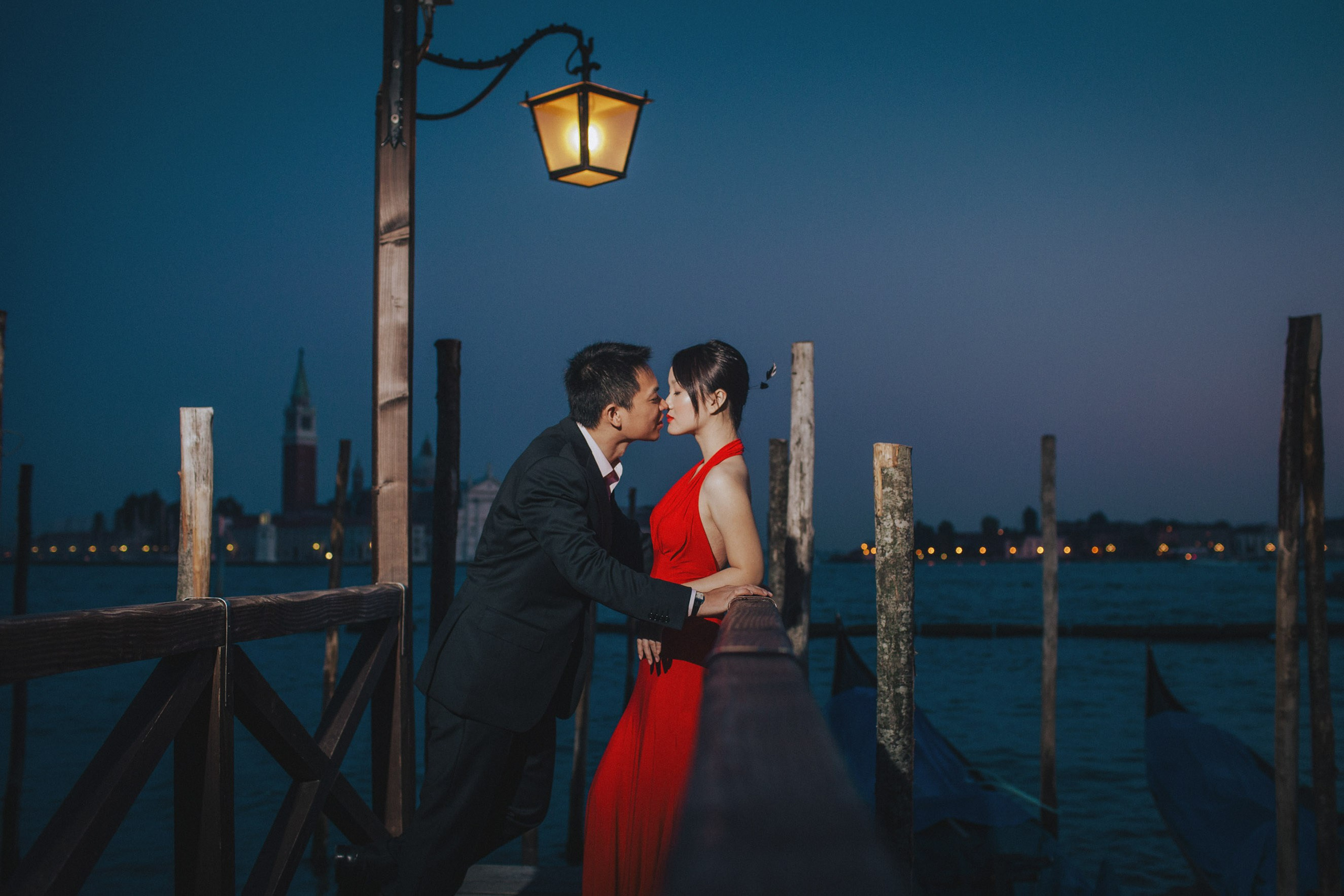 A Thai man leans in for a kiss as his red dress wearing woman leans back against a railing above the gondola's during their sexy Love Story portrait session in Venice.