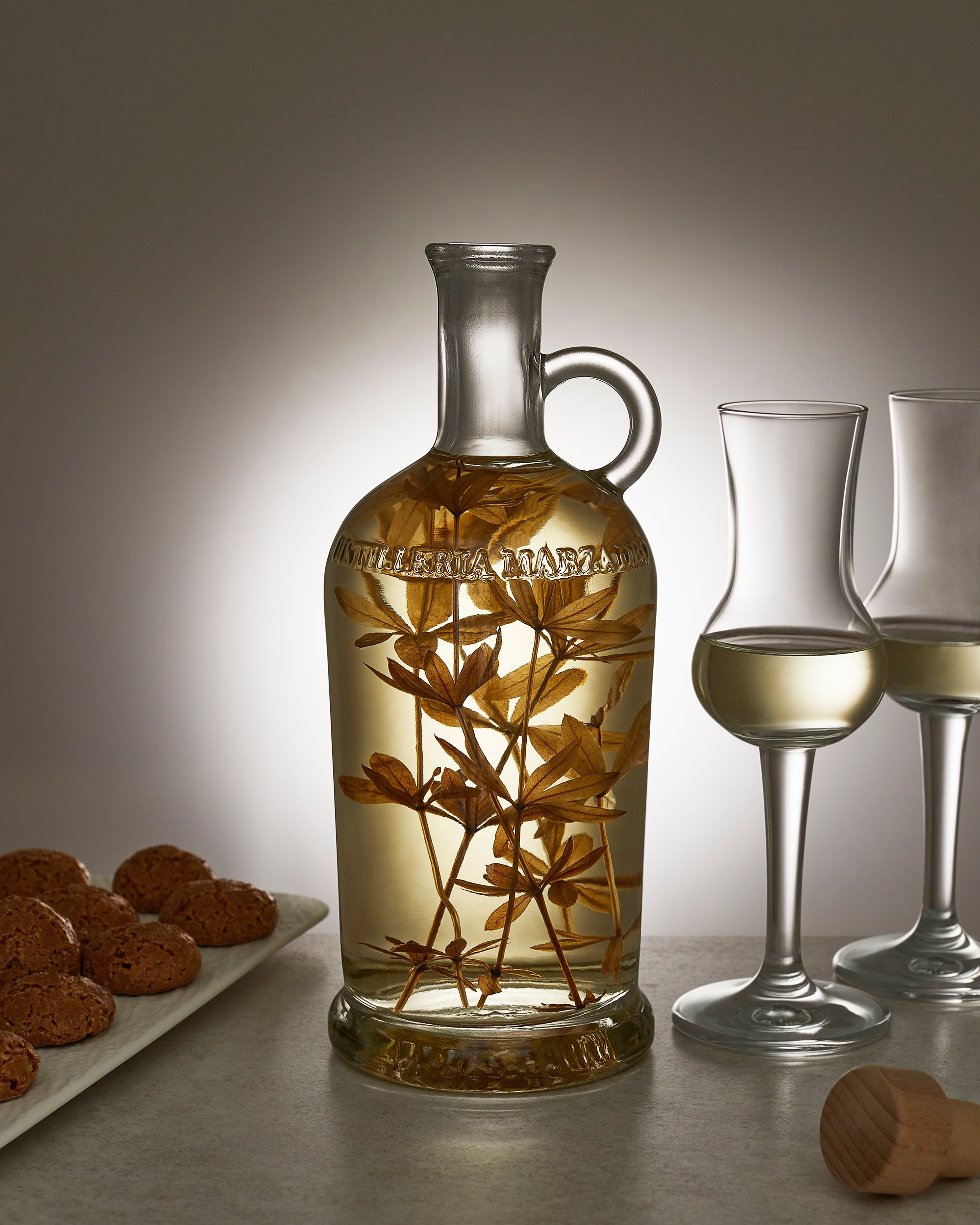 Photographer Roman Djuzev - A bottle of grappa with asperula grass, two glasses of grappa and a light snack of almond cookies on a stone table.