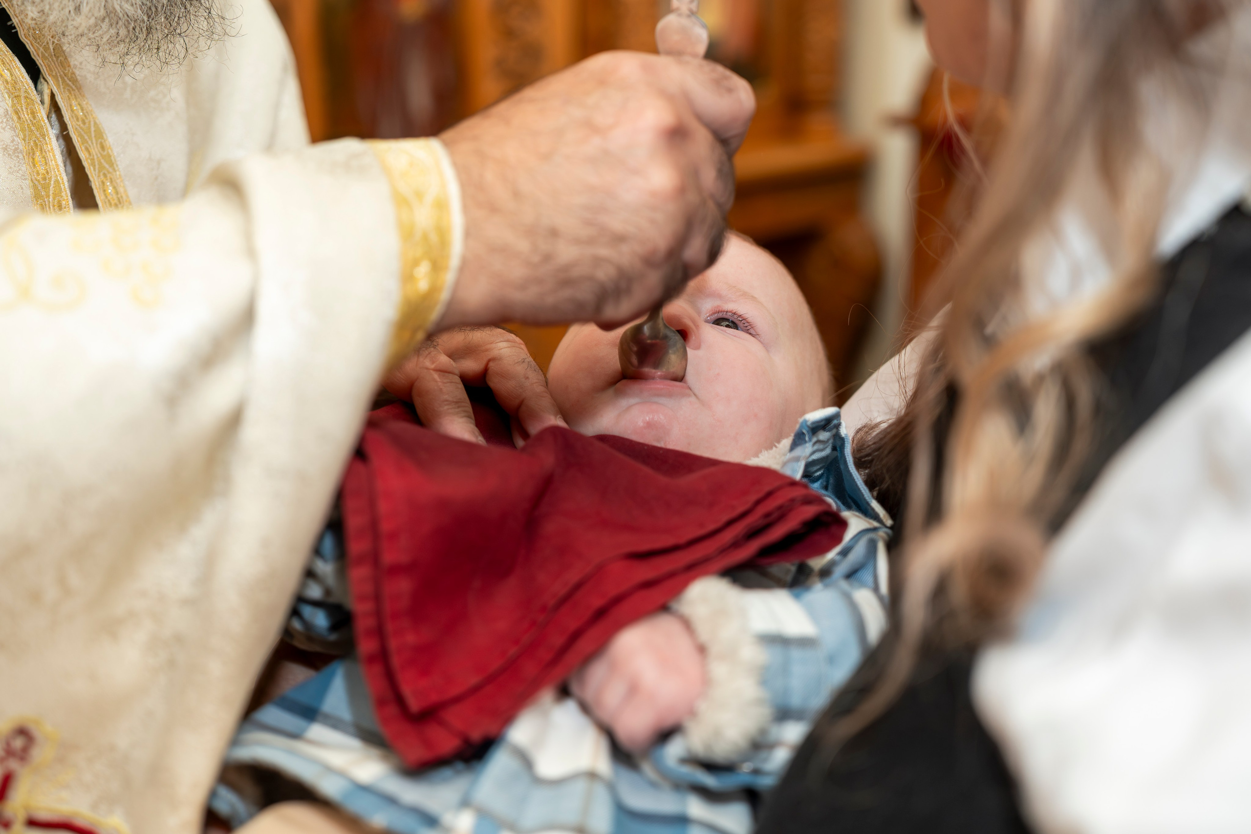 Priest administering baptismal spoon to baby held by adult during baptism ceremony.
