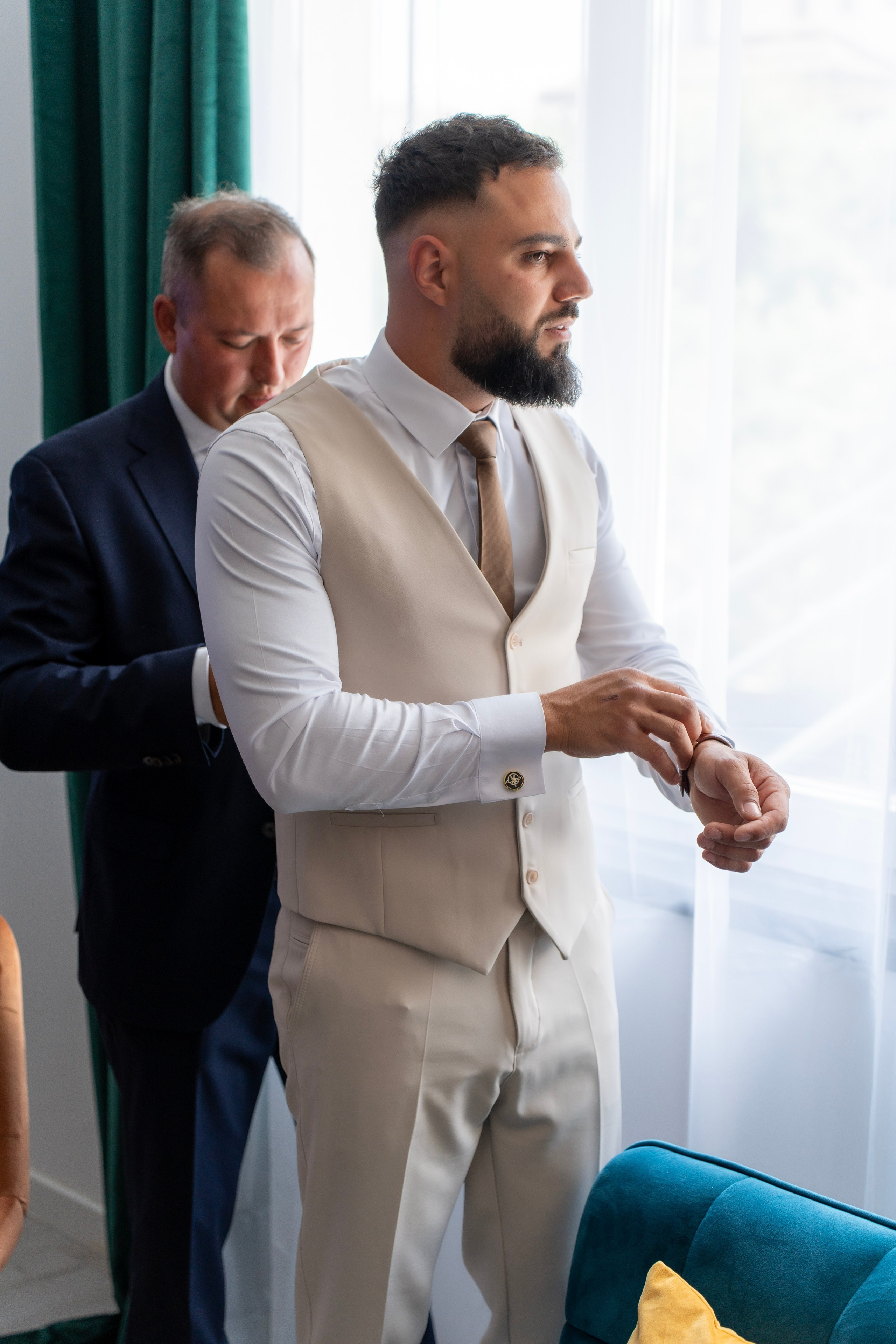 Man in beige vest adjusts watch while another man helps with vest near bright window.