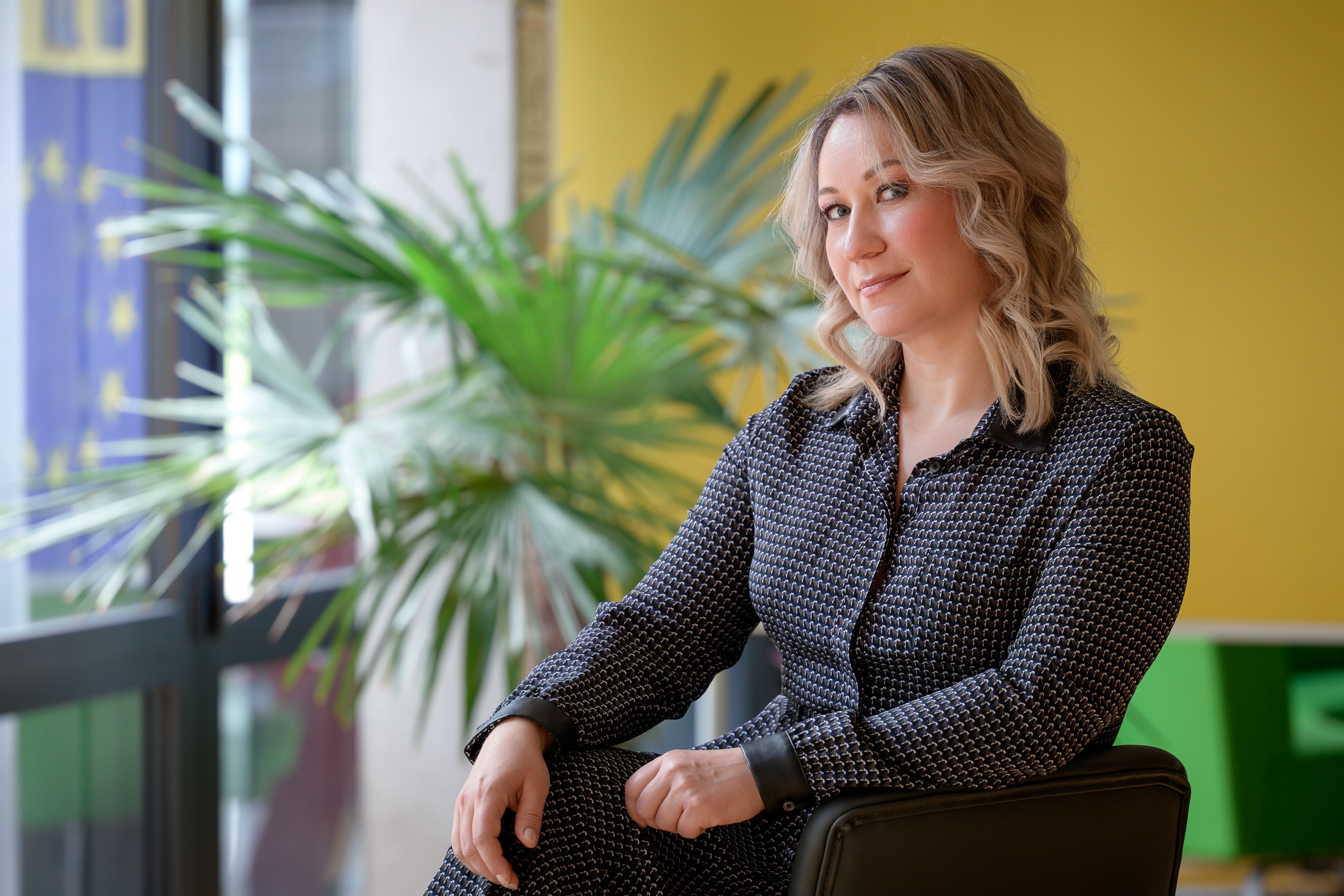 Ein Business-Porträt einer jungen Frau in einem dunklen, bunten Kleid vor einem gelben Hintergrund mit einer Palme.
