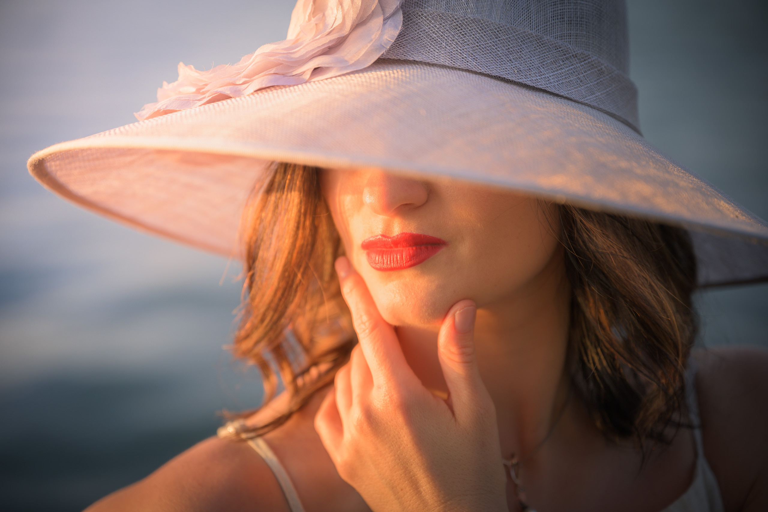 Eine junge Brünette in einem beigen Overall und einem puderfarbenen Hut fängt mit ihrem Gesicht die letzten Strahlen des sommerlichen Sonnenuntergangs ein.