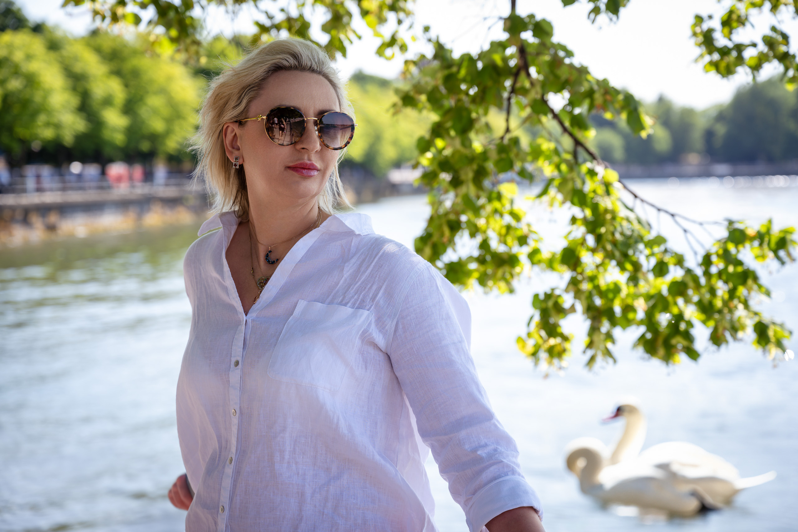 Eine 40-jährige Blondine in einem weißen Leinenhemd posiert im Schatten vor einem See mit Schwänen.