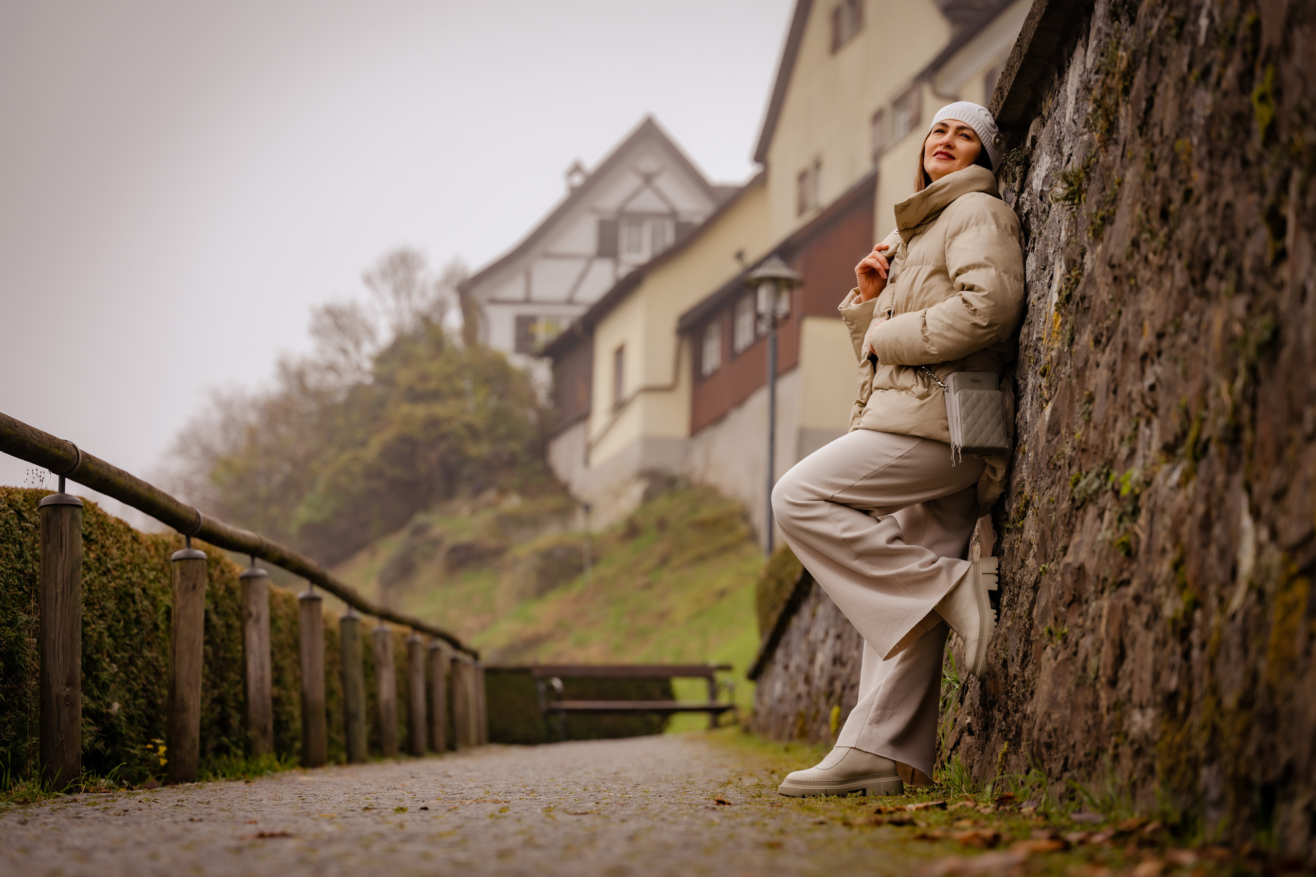 Eine geheimnisvolle Frau posiert an einem nebligen Herbsttag vor einem alten Kloster, lehnend an einer Steinmauer.