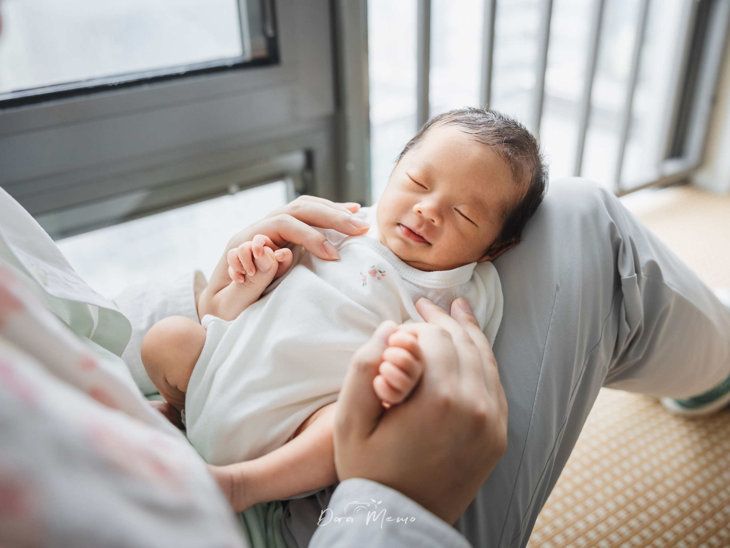 Father sitting by the window holding his sleeping newborn, quiet guardianship in soft light