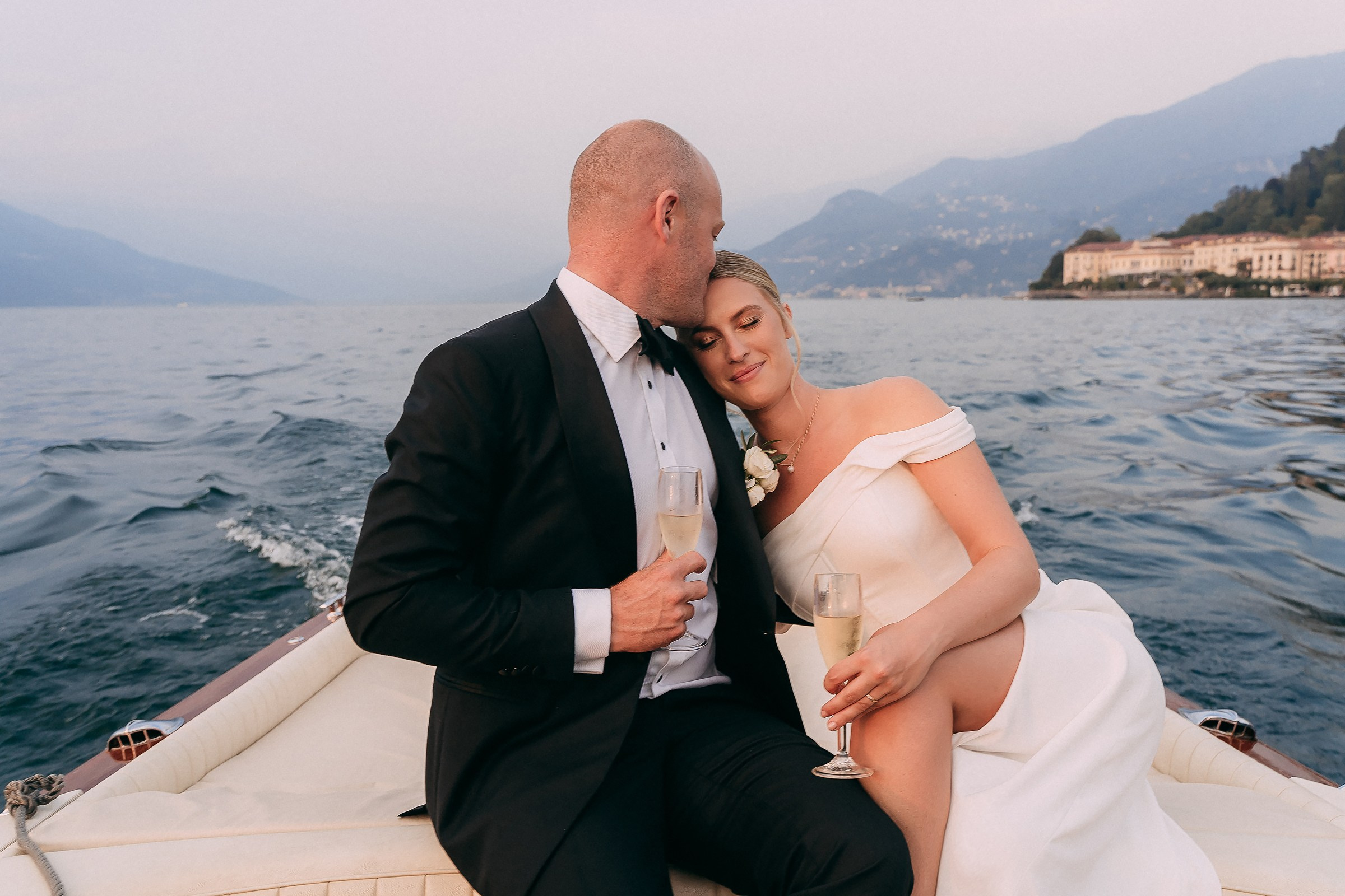 A bride and groom seated closely on a boat, enjoying a serene moment with champagne, the bride resting her head on the groom’s shoulder as they glide across the water.