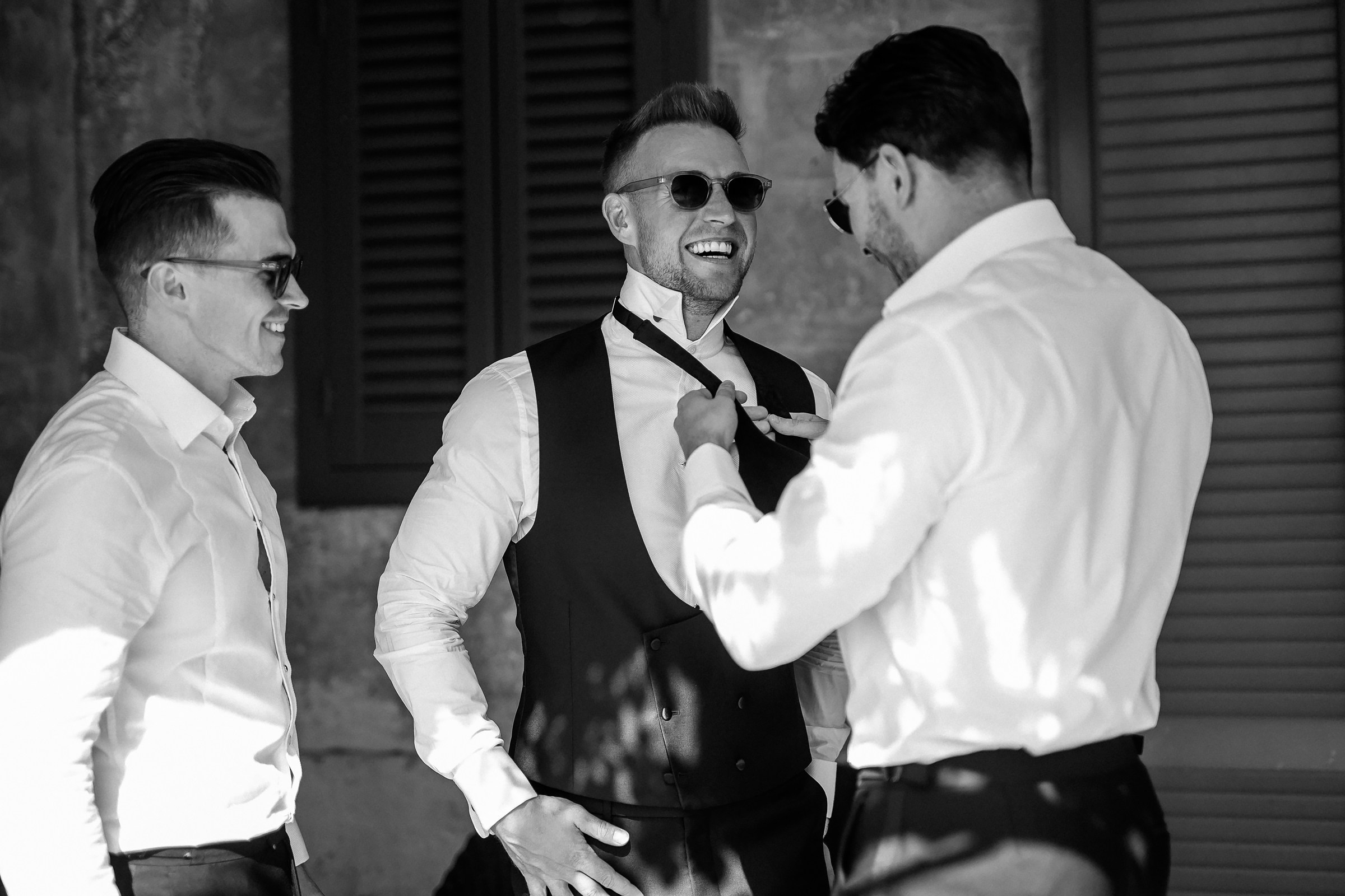 Laughter fills the air as the groom’s friends help him perfect his bowtie, a casual and heartwarming pre-wedding moment captured in black and white.
