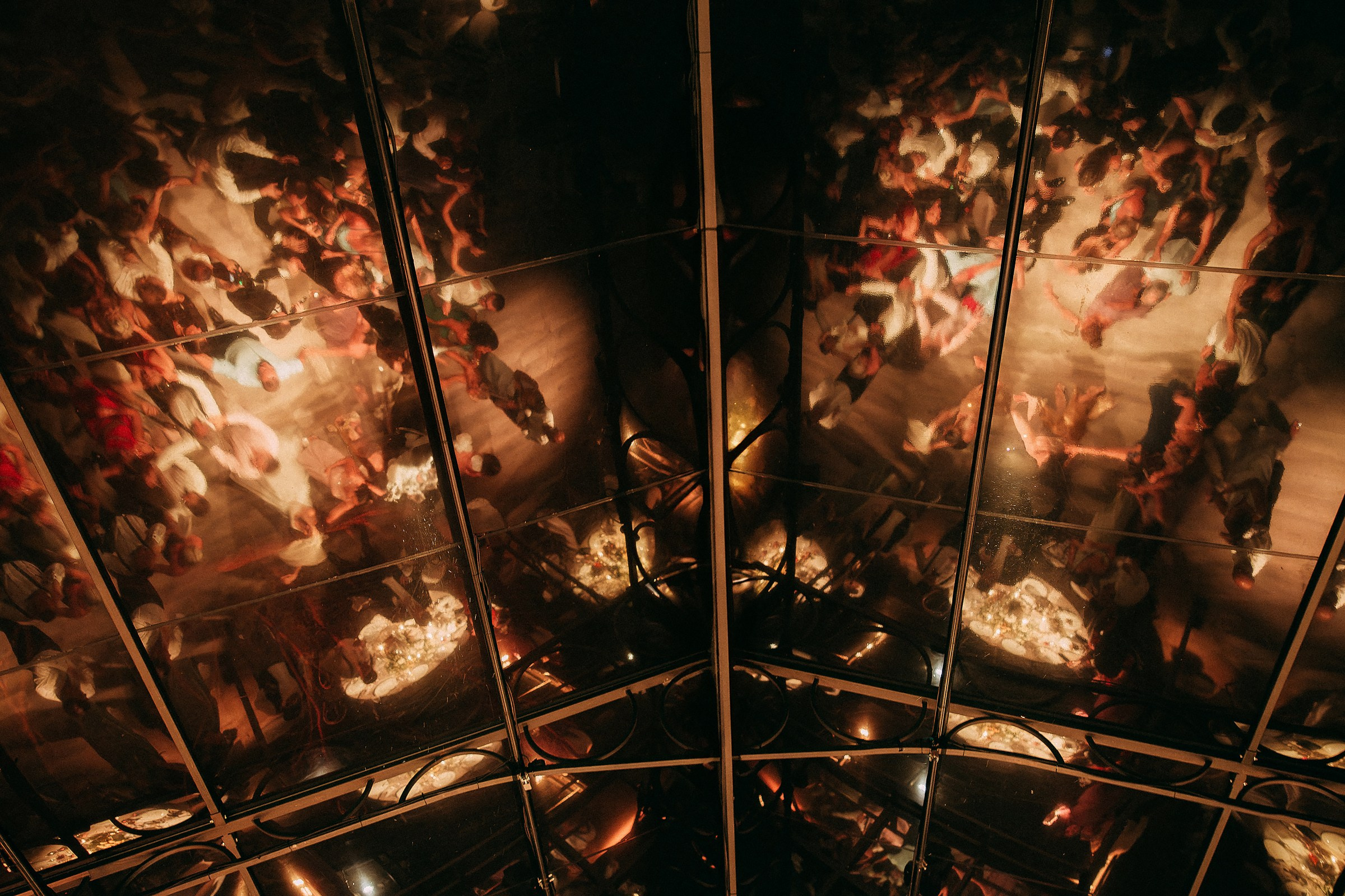Reflection of dancing guests in glass ceiling, capturing vibrant wedding energy.