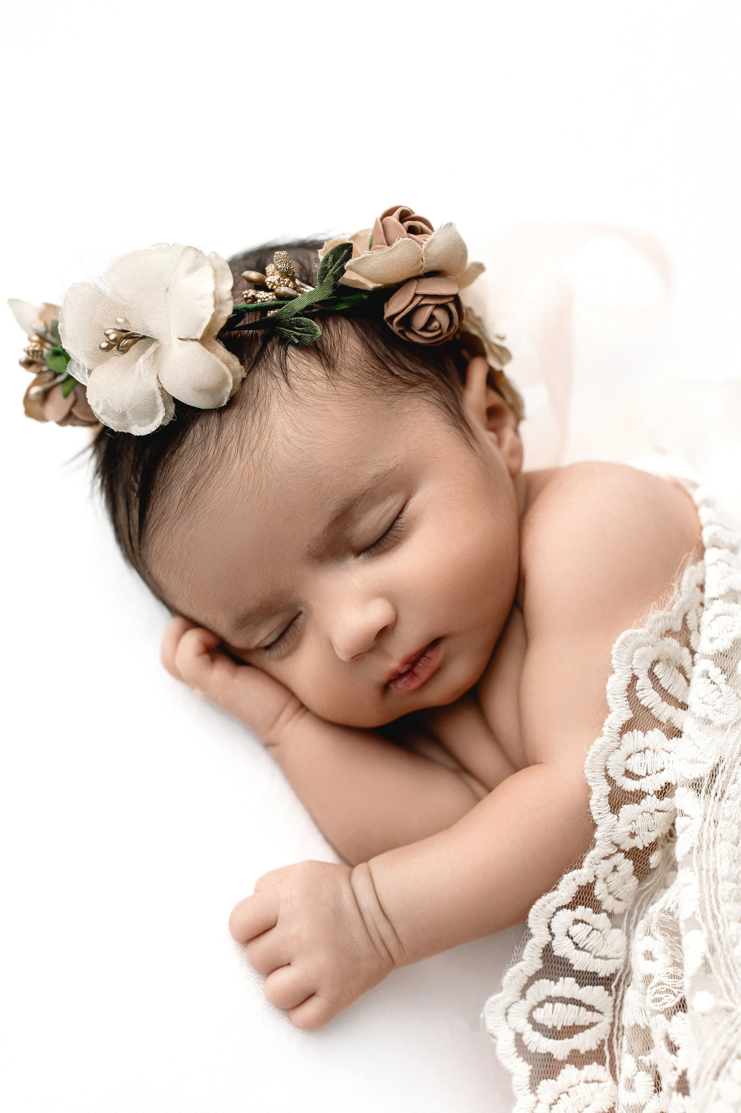 Newborn baby girl sleeping on a white blanket, wearing a floral headband and lace wrap — elegant and natural newborn portrait with soft tones.