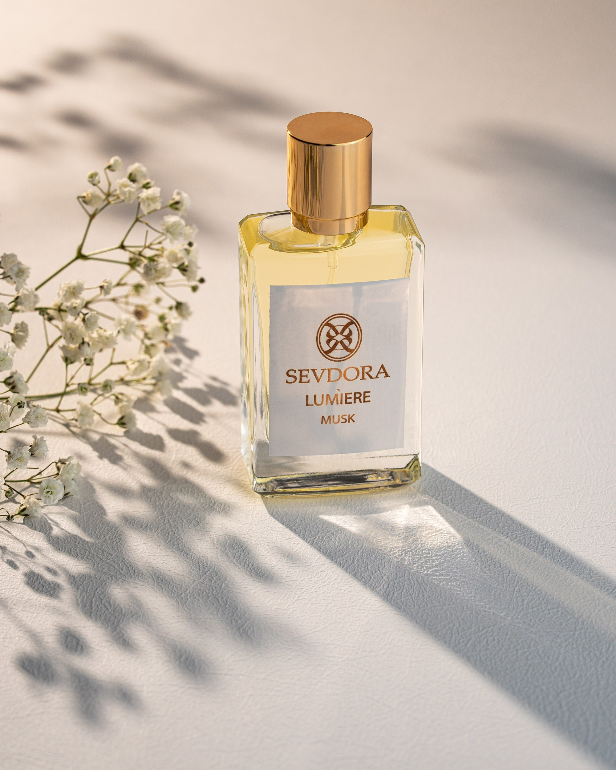 Açıq rəngli səth üzərində, günəş işığı və kölgələr altında duran, "SEVDORA LUMIERE MUSK" yazısı olan şüşə ətir butulkası. Butulkanın yanında zərif ağ çiçəklərdən bir budaq var.