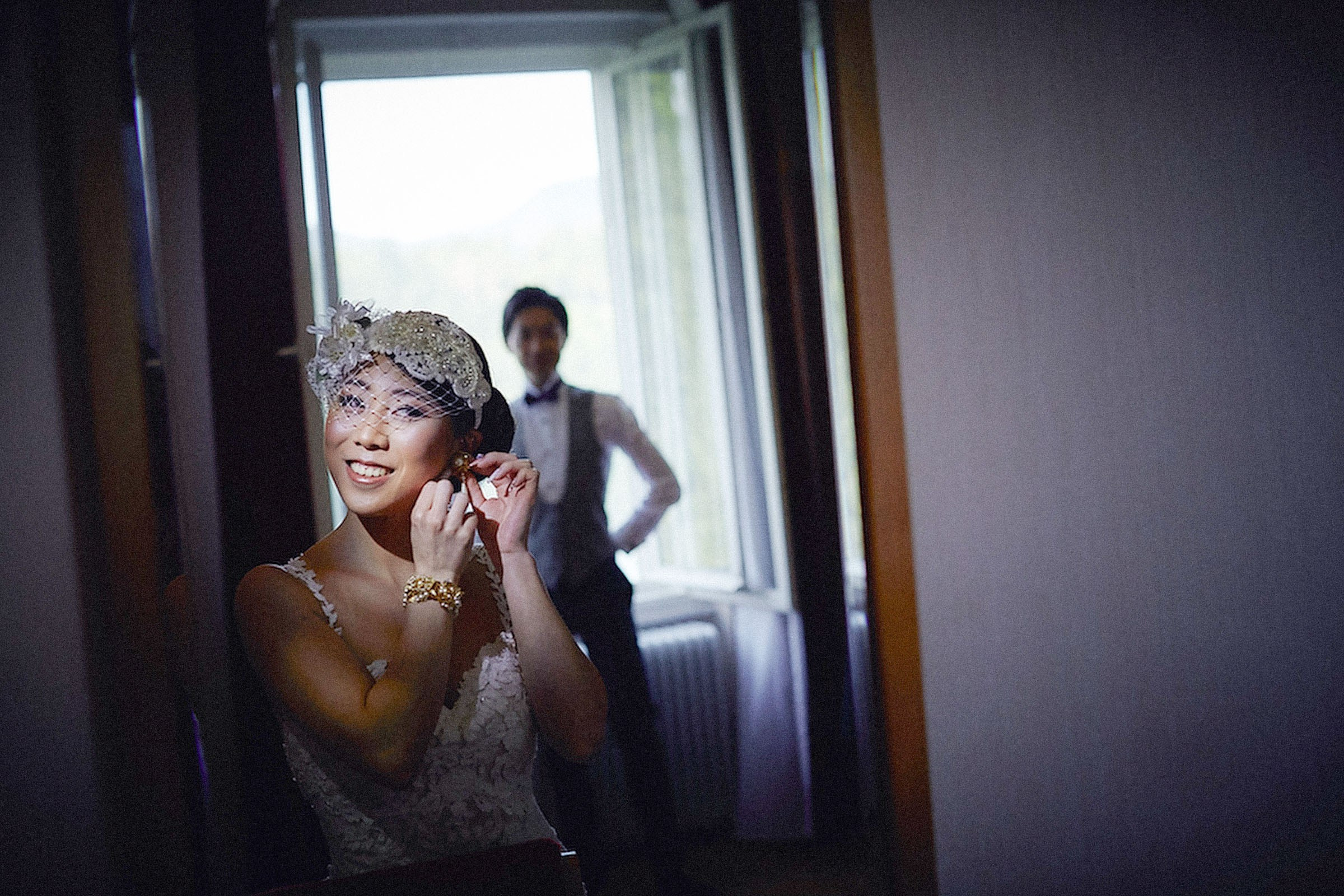 A bride wearing a birdcage veil puts on her earrings as her groom stands behind her admiring her wedding attire.