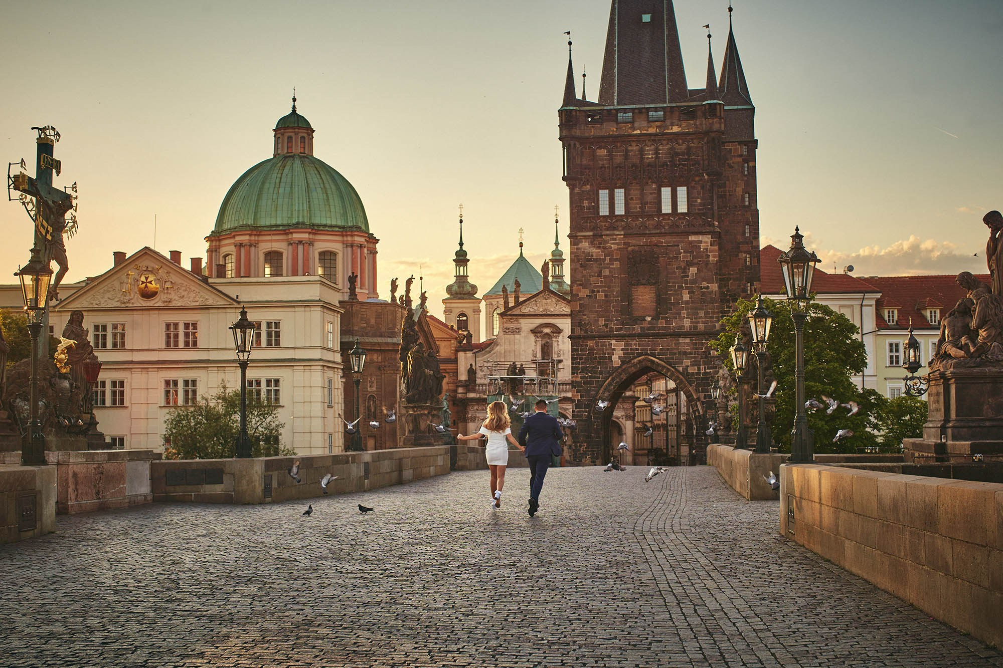 Blonde fiancée in flowing white dress running with fiancé across empty Charles Bridge as pigeons soar.