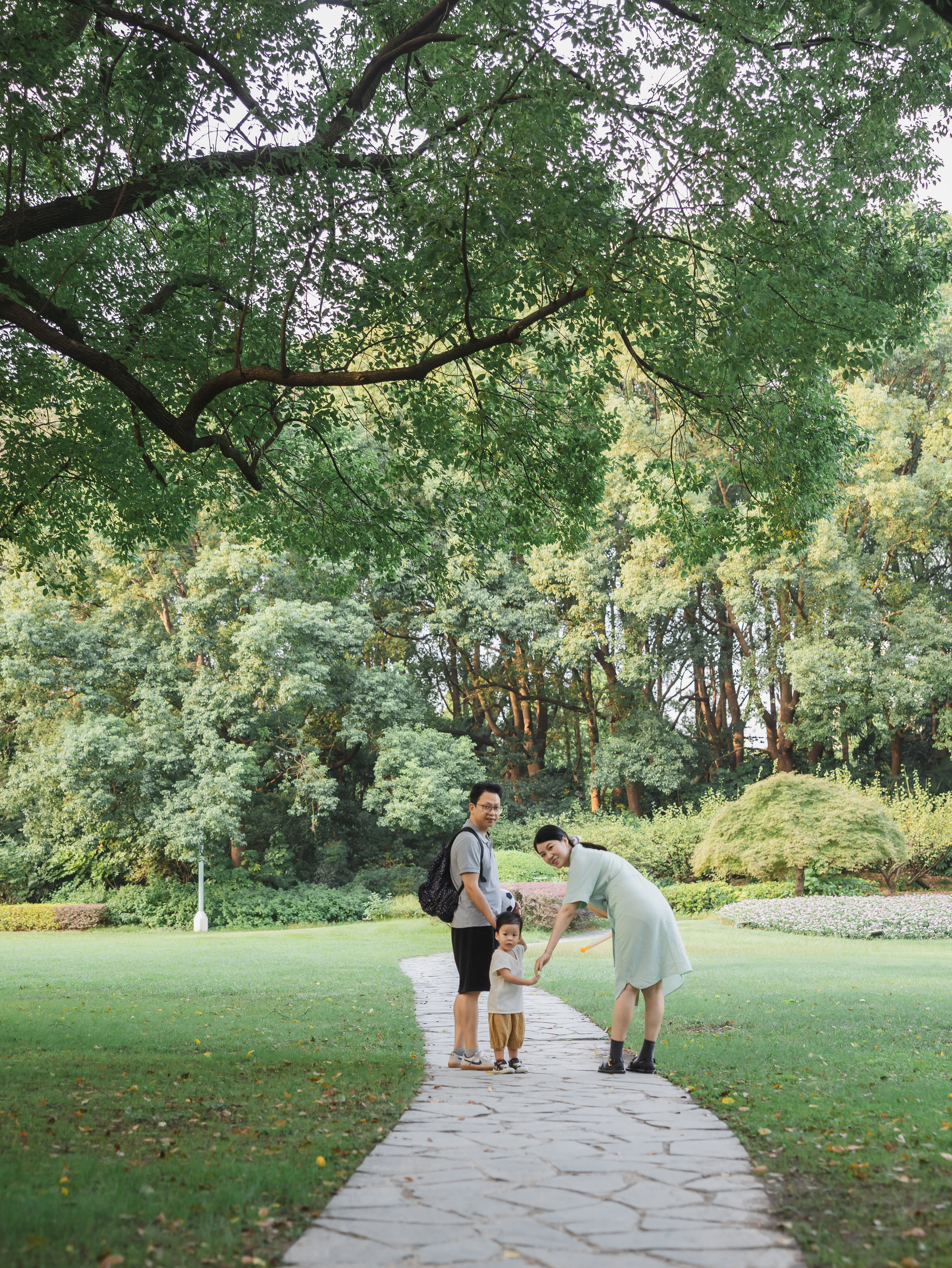 A family of three walks away along a stone path under large trees in a peaceful Shanghai park.